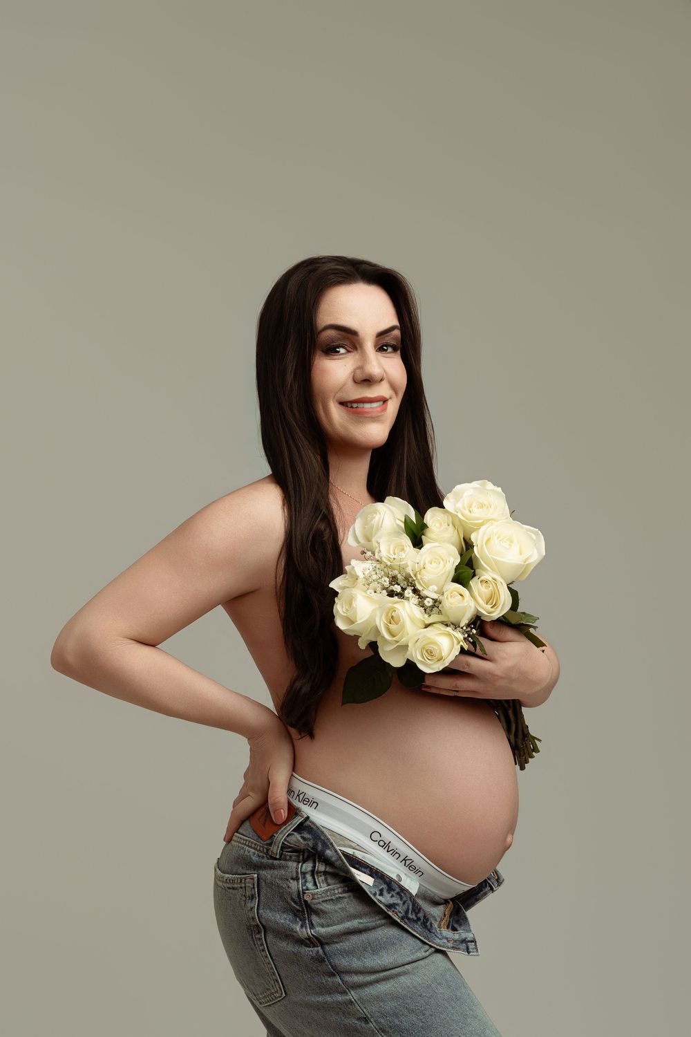 Pregnant woman holding white roses bouquet, wearing open jeans, smiling against gray background.