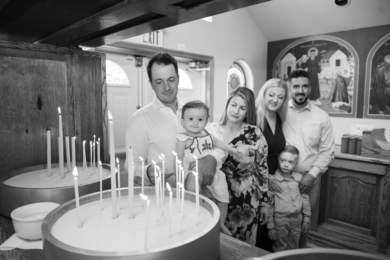 Family gathered around baptismal font with candles in Orthodox church during christening ceremony.