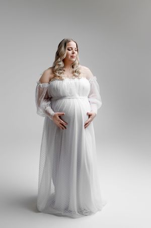 A pregnant woman poses in an ethereal white off-shoulder maternity gown against a light gray studio backdrop.