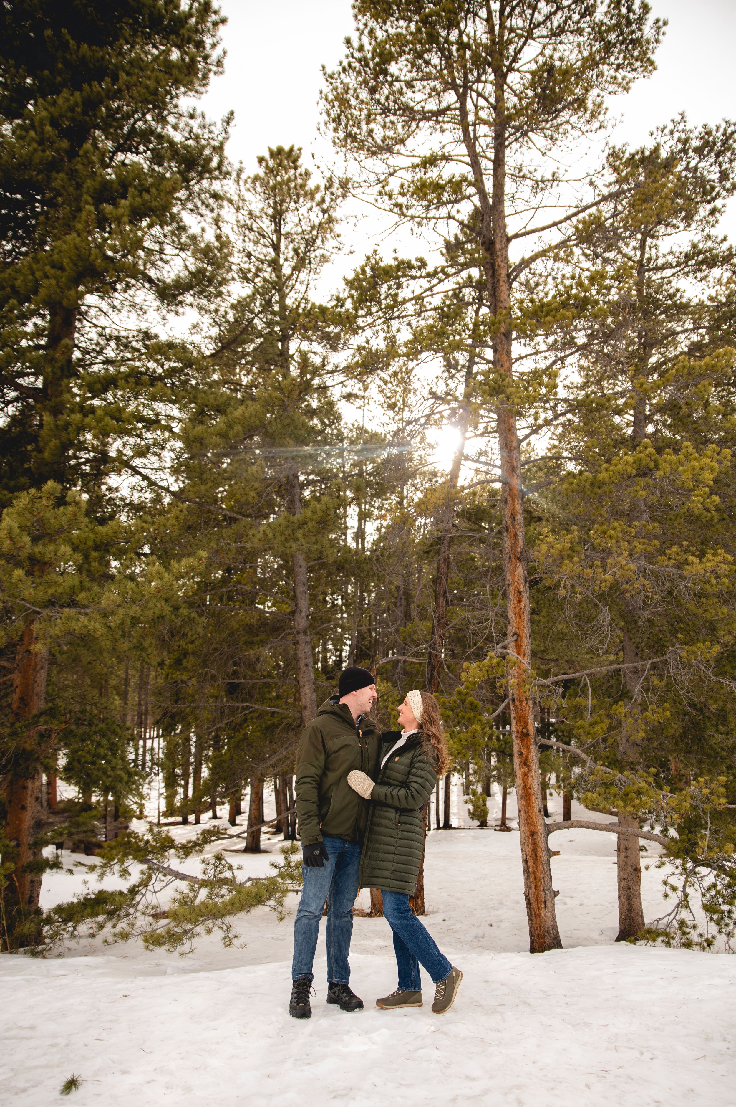 Couple embracing and smiling at each other in a snowy pine forest during winter, wearing warm coats and hats.