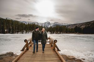 Couple walking hand-in-hand on a wooden dock overlooking a frozen lake with snowy mountains in winter.
