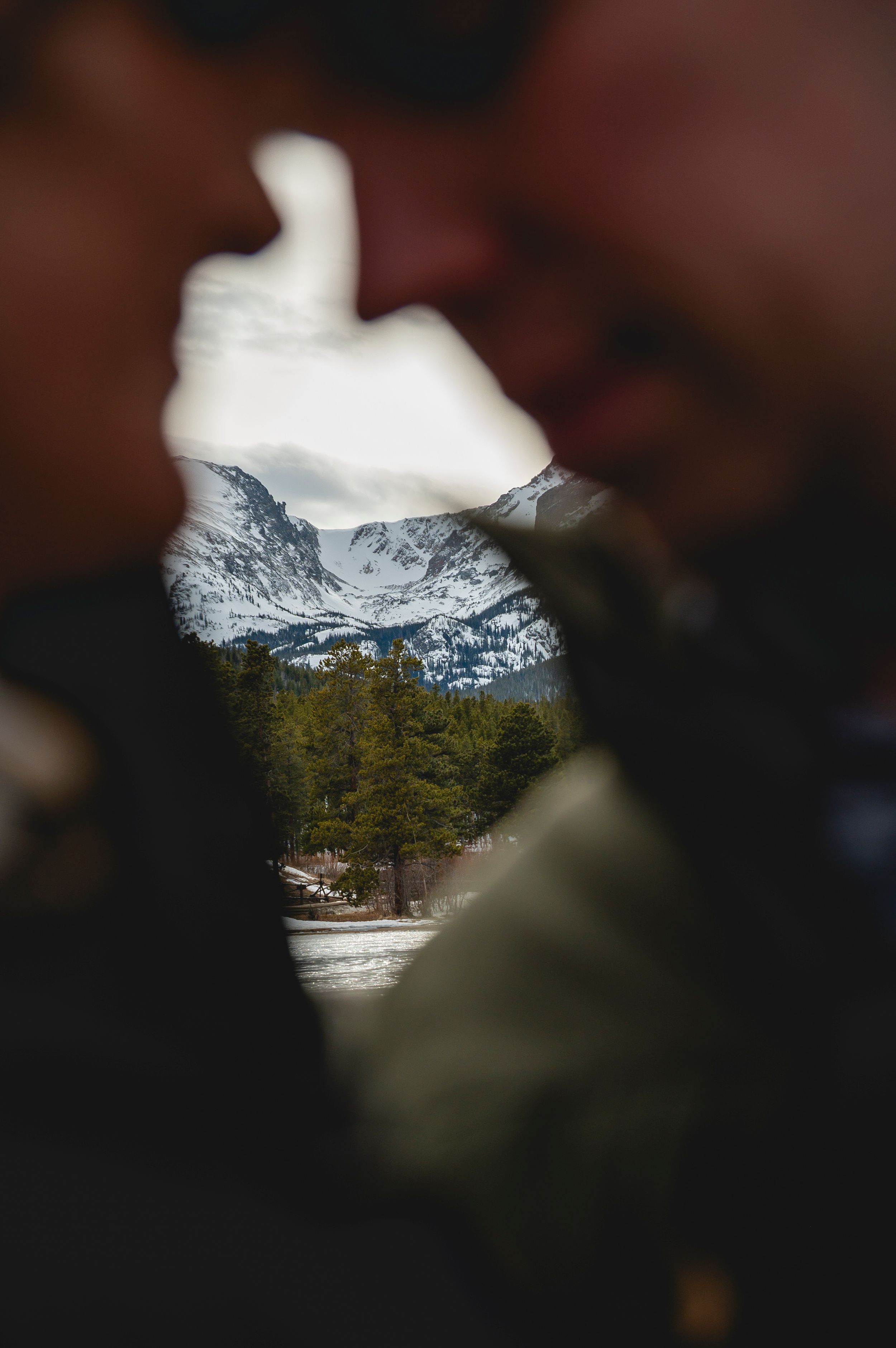 Couple kissing framed by snowy mountain peaks and evergreen trees visible through the gap between their faces.
