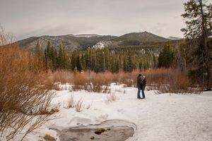 Couple standing in a snowy winter landscape with mountain forest backdrop.
