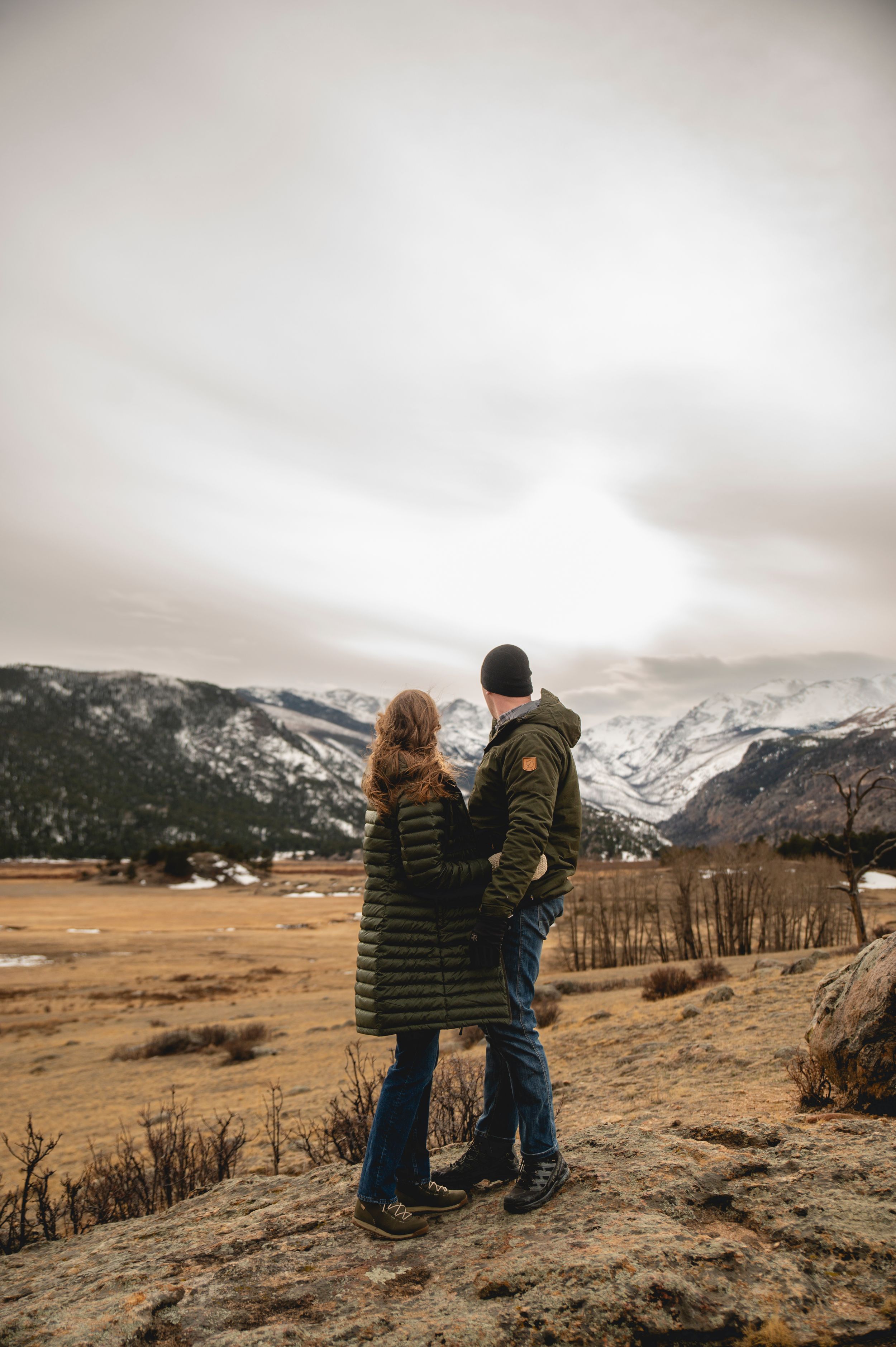 Couple in green winter coats embracing on rocky overlook with snow-capped mountains and overcast sky.