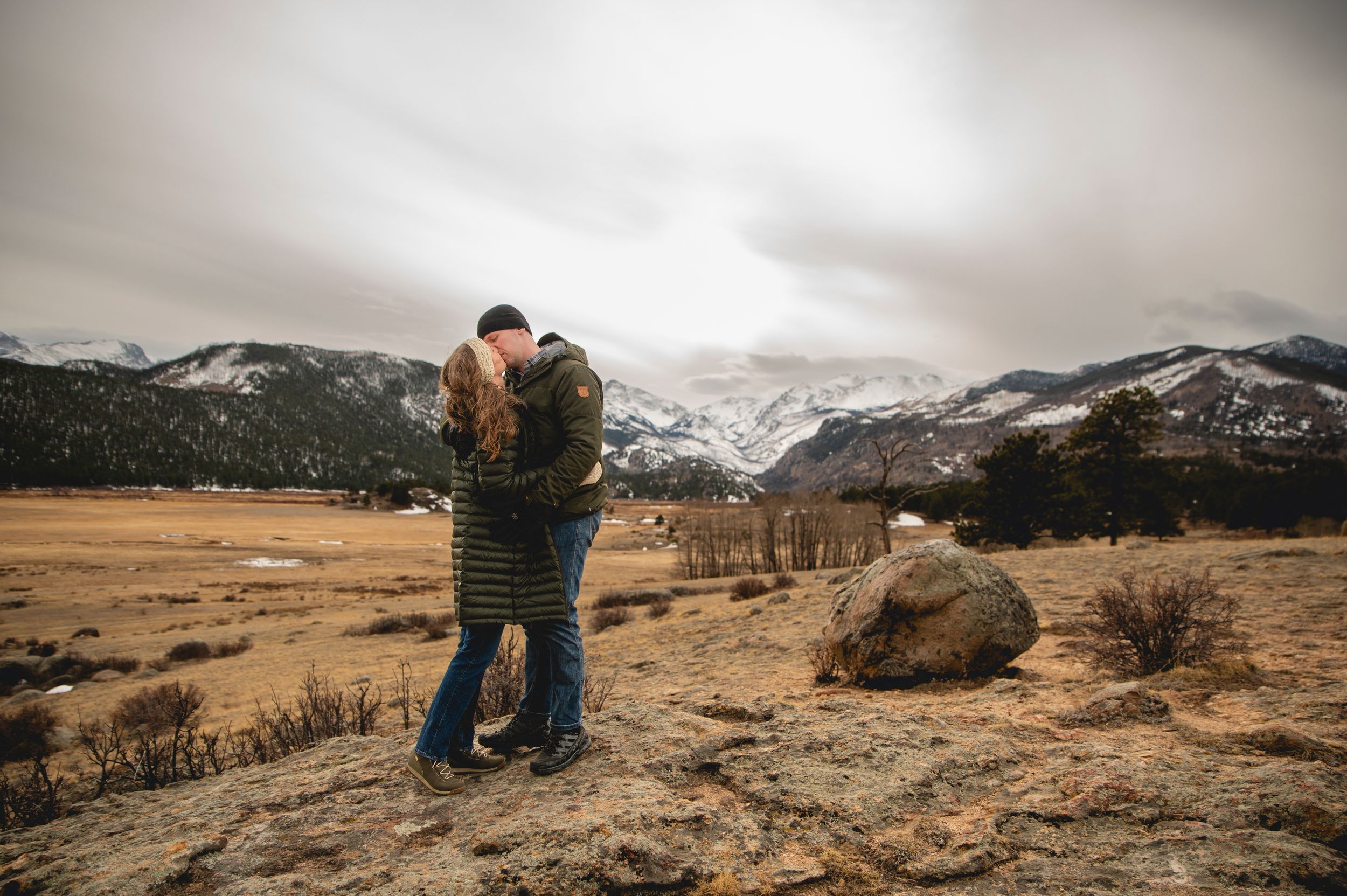 Couple embracing on rocky terrain with snow-capped mountains and overcast sky in background.