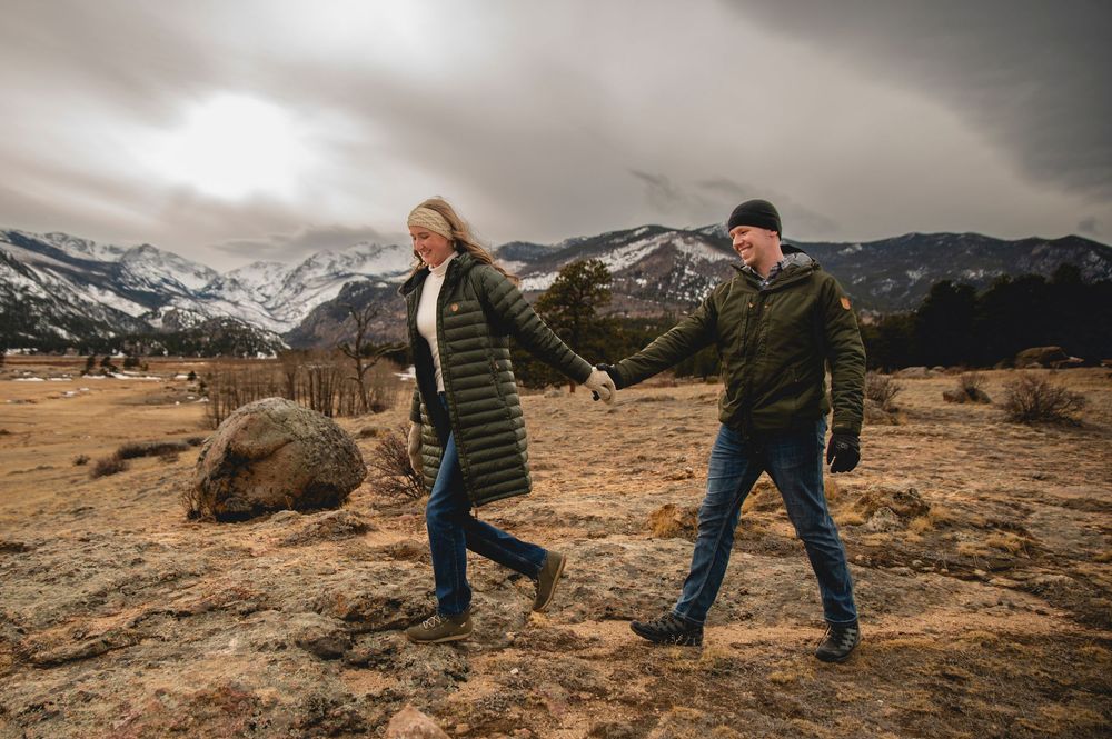 Couple holding hands walking through a rocky mountain meadow with snow-capped peaks and dramatic cloudy skies.
