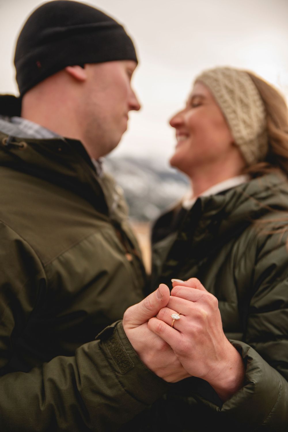 Engaged couple in green jackets holding hands outdoors, showing off diamond engagement ring, smiling at each other.