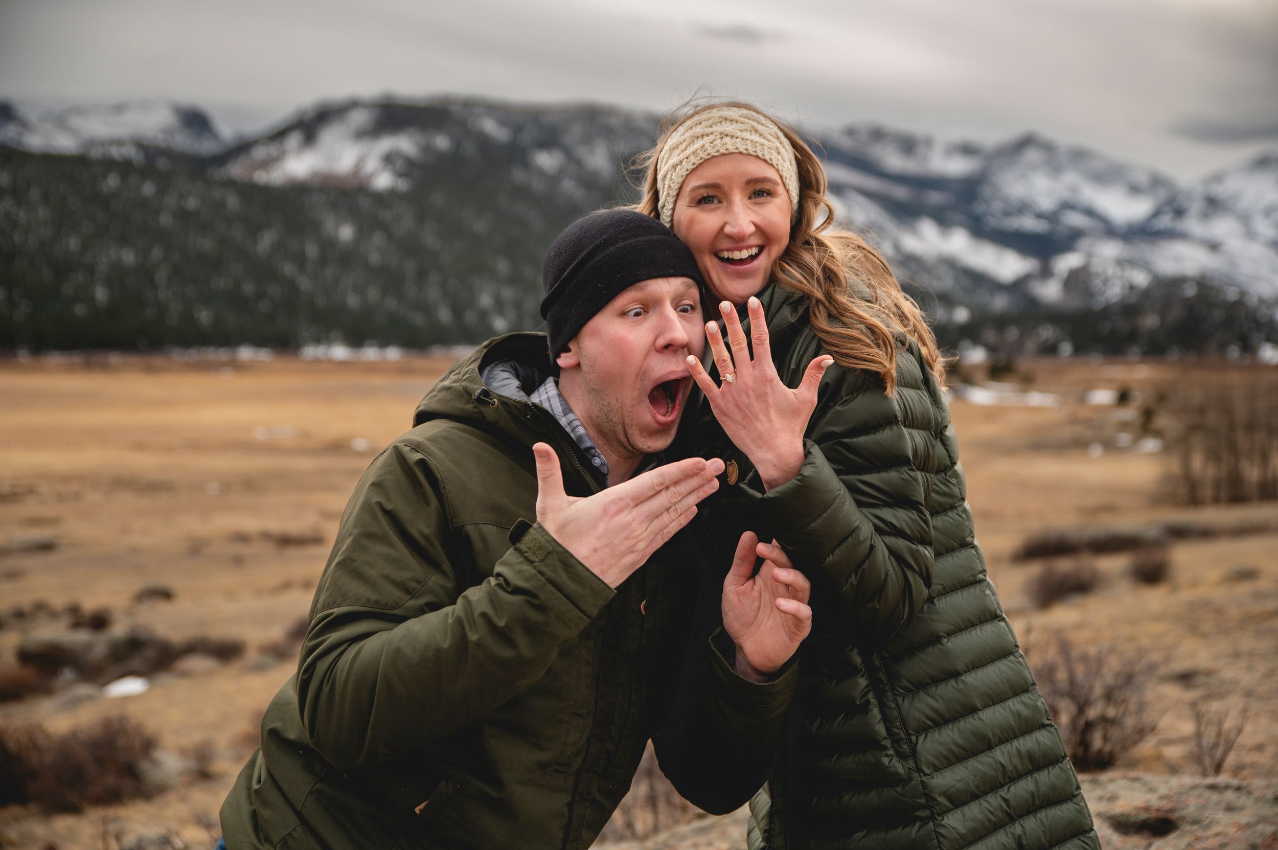 Excited couple showing off engagement ring outdoors with mountain backdrop in winter landscape.