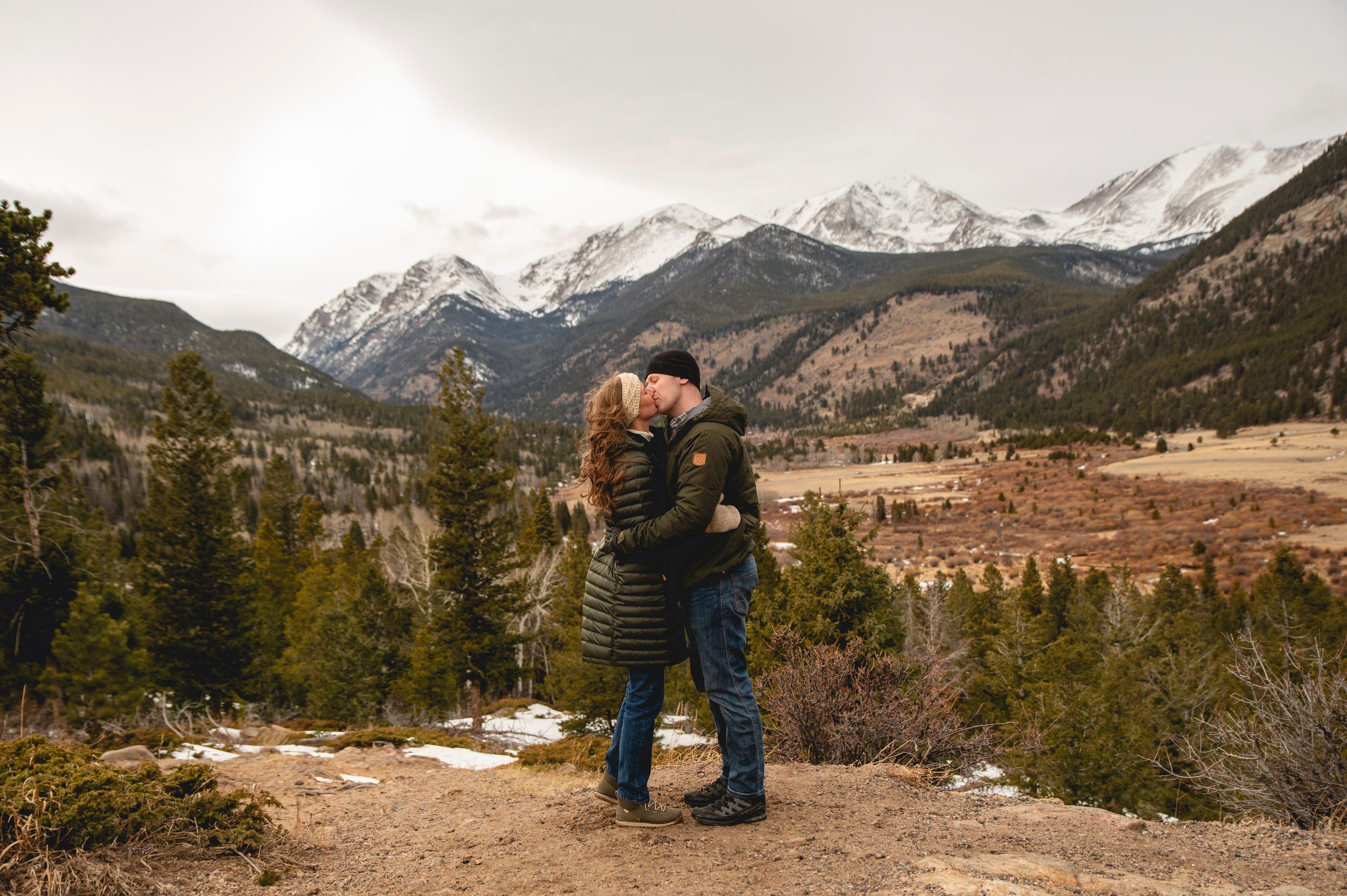 Couple embracing on a mountain overlook with snow-capped peaks and evergreen forest in Rocky Mountain National Park.