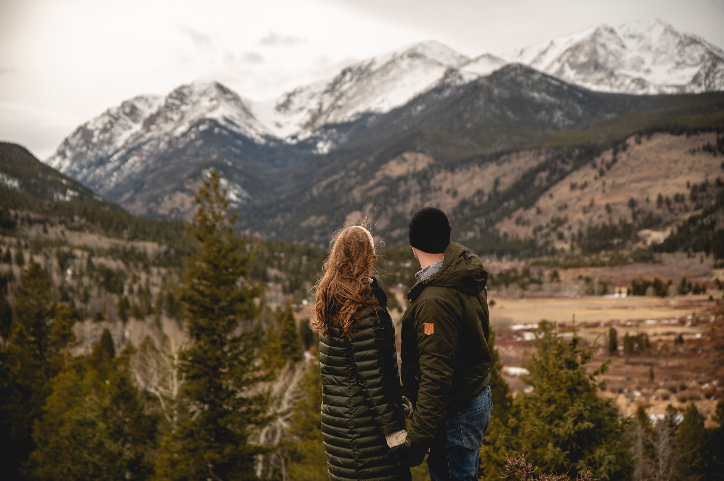 Couple holding hands admiring snow-capped Rocky Mountain peaks from a scenic overlook surrounded by pine trees.