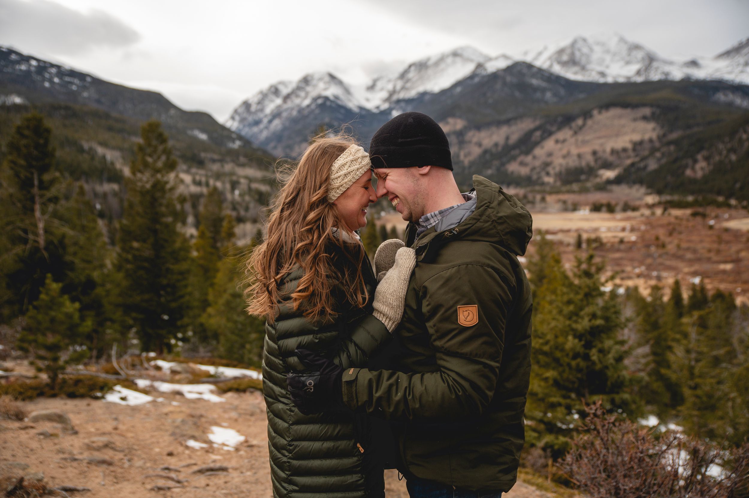 Couple embracing and smiling at each other with snow-capped Rocky Mountains and pine trees in the background.