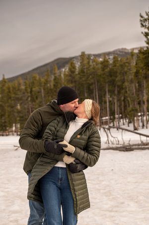 Couple kissing in snowy mountain landscape wearing matching olive puffer jackets and winter accessories.