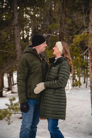 Couple in matching green winter jackets embracing in a snowy forest setting, smiling at each other.
