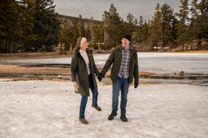 Couple holding hands and smiling on a snowy frozen lake surrounded by pine trees in winter.