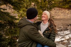 A happy couple embraces outdoors in winter, surrounded by snow-dusted pine trees, both wearing cozy jackets and knit hats.