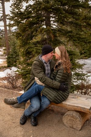 Couple in matching olive puffer jackets sitting close on a rustic log bench, surrounded by snowy pine trees.