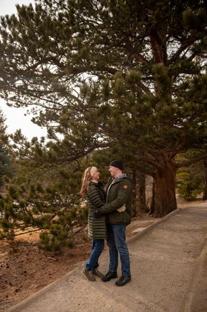 Couple embracing on a scenic path surrounded by tall pine trees during a winter outdoor engagement photo session.