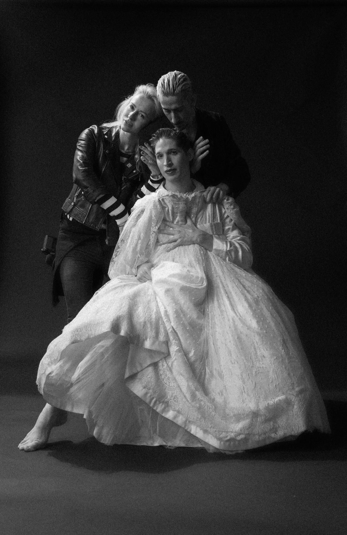 Black and white photo of three theatrical performers, woman in white gown seated with two costumed figures behind her.