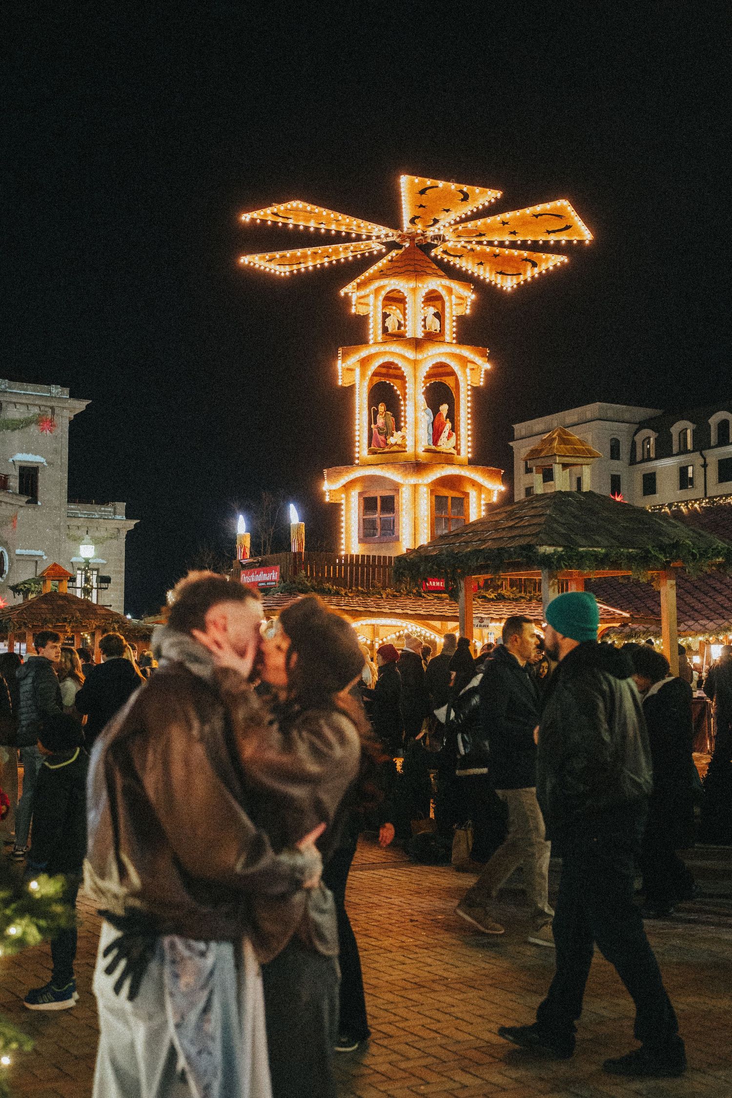 Couple kissing at a festive Christmas market at night with a lit pyramid decoration in the background.