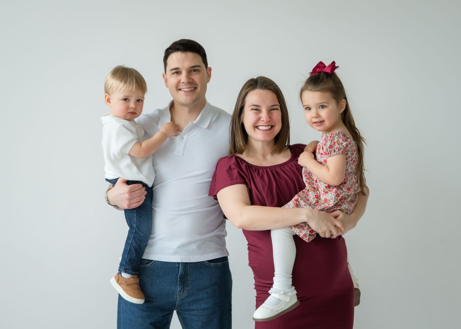 Happy family of four posing for a portrait, parents holding two young children, smiling against a white background.
