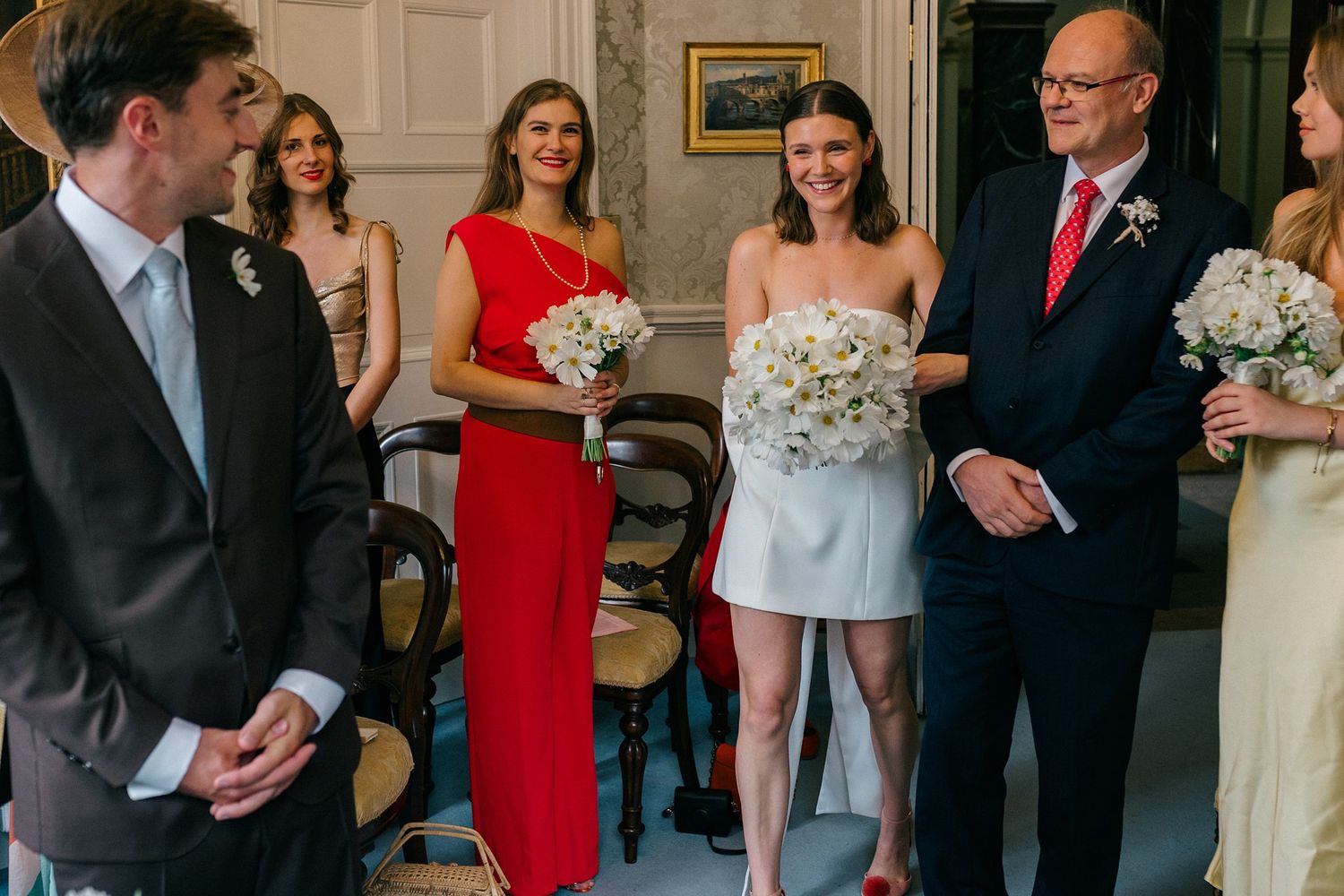 Bride in short white dress walks down aisle with father, flanked by bridesmaids in red and nude gowns holding white bouquets.