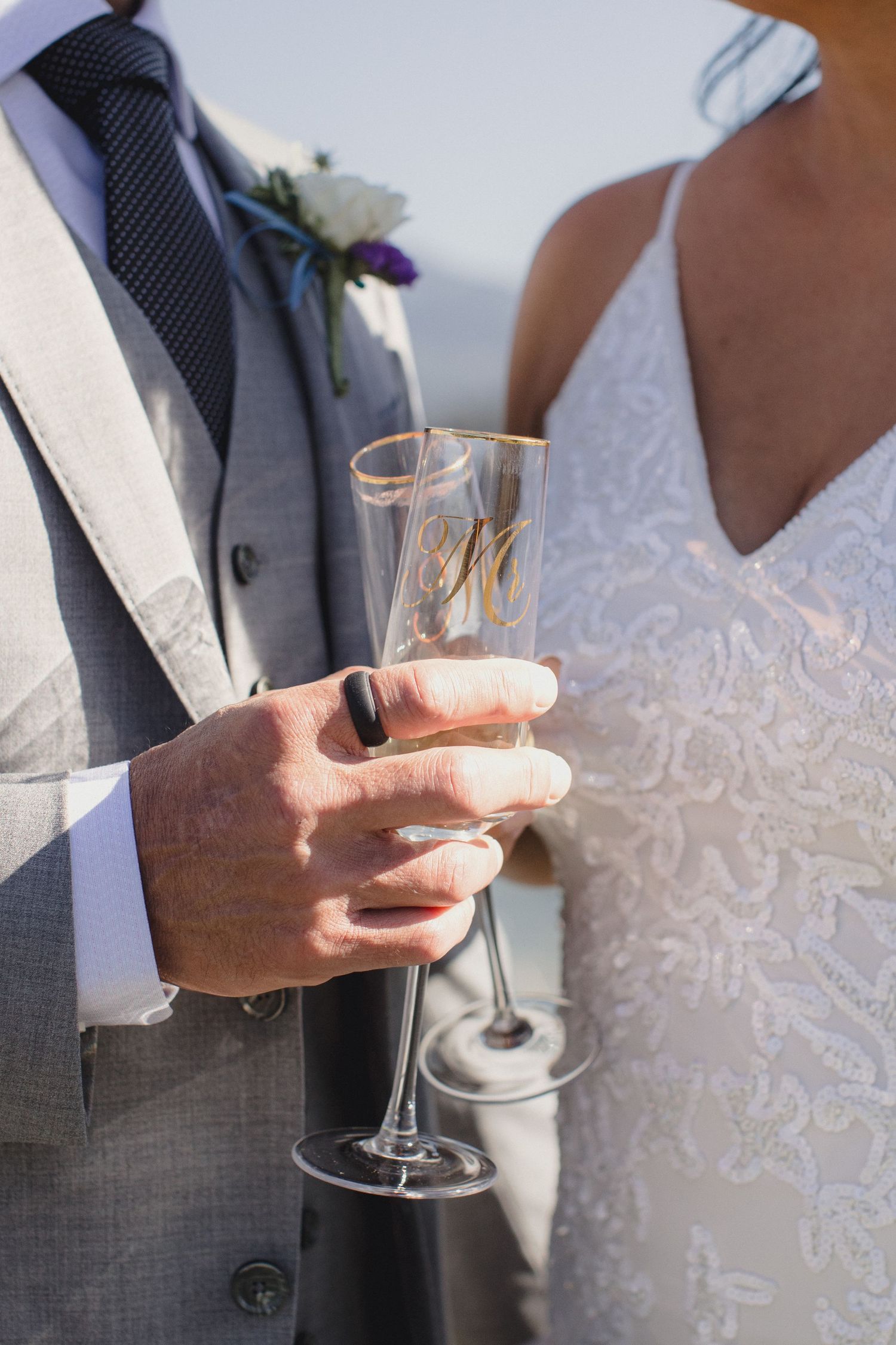 Bride and groom holding personalized champagne flutes at wedding, groom in gray suit with boutonniere.