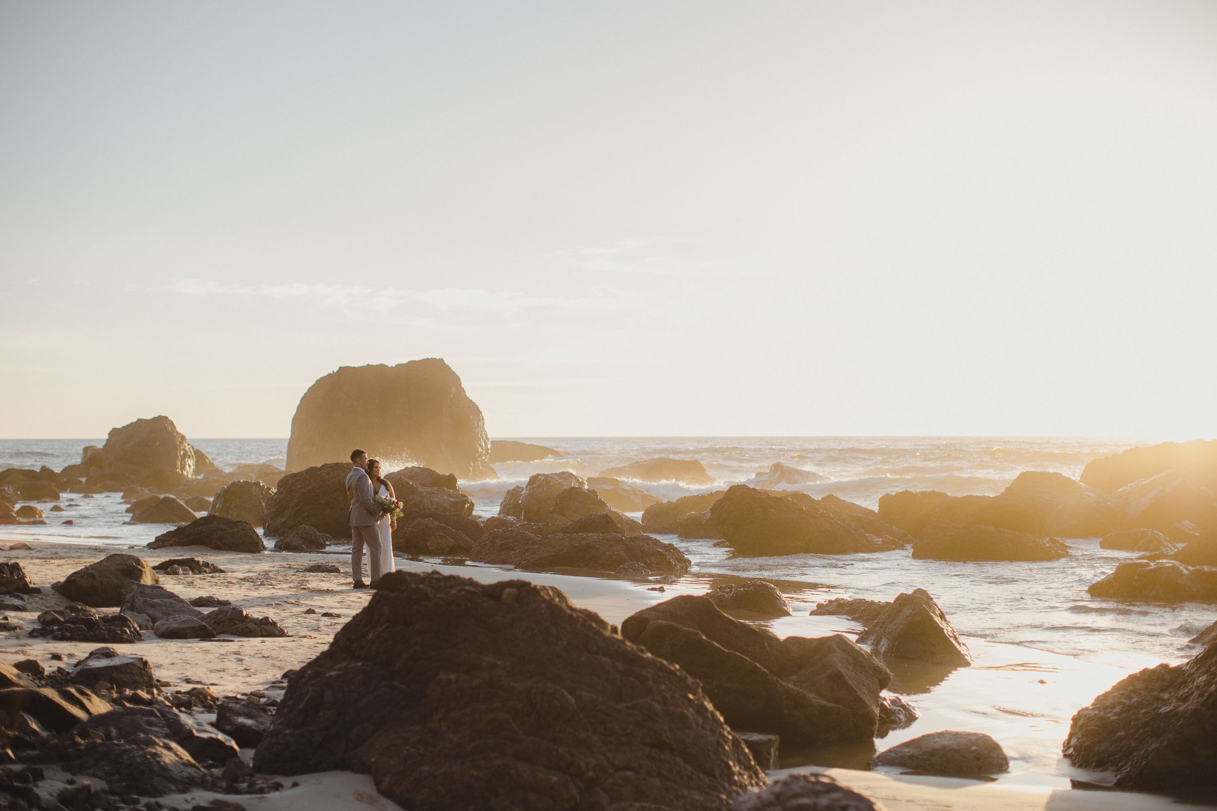 Couple embracing on rocky beach at golden sunset with ocean waves and large boulders in background.