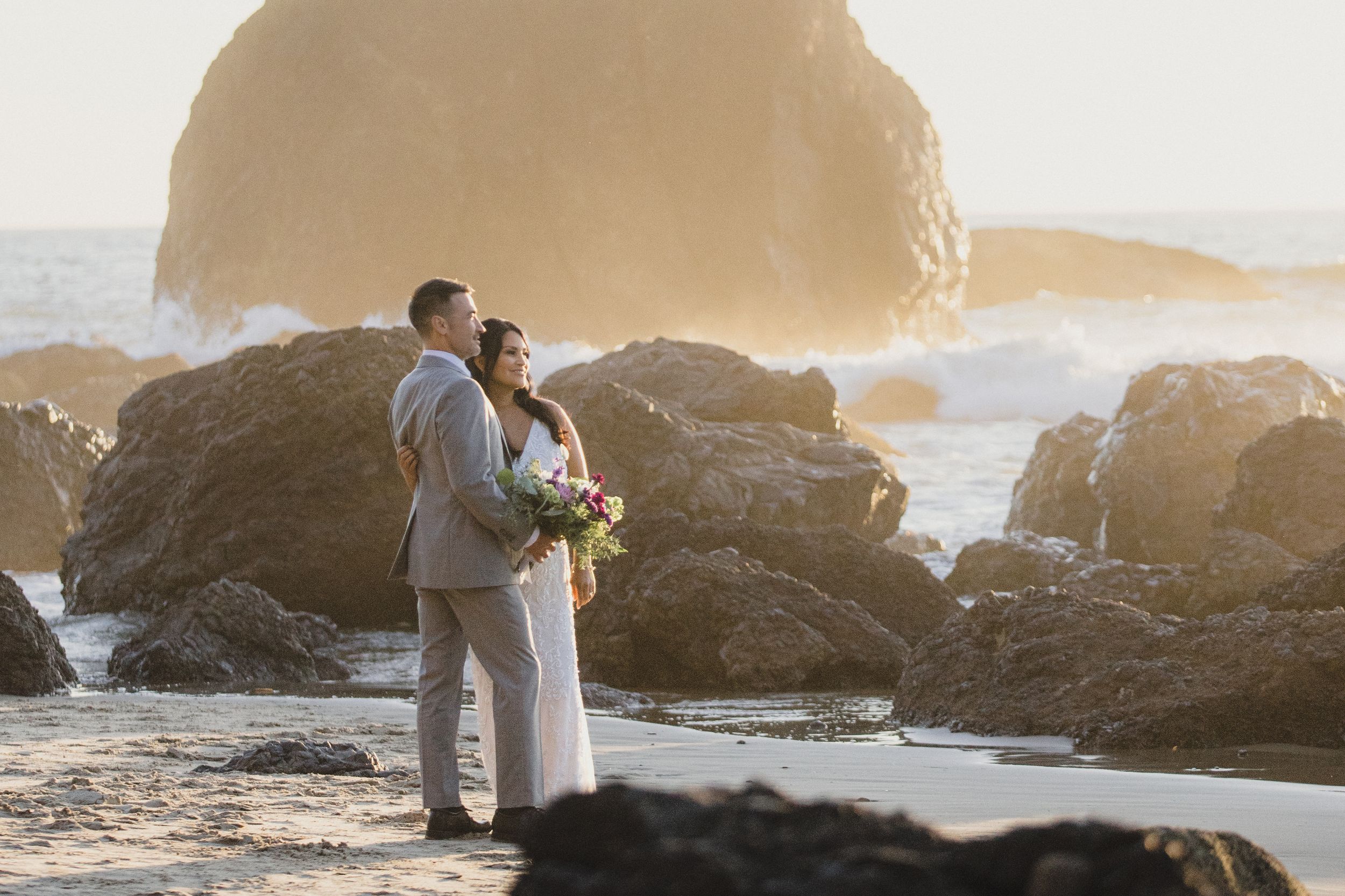 Couple in wedding attire standing on rocky beach at sunset with ocean waves and large boulders in background.