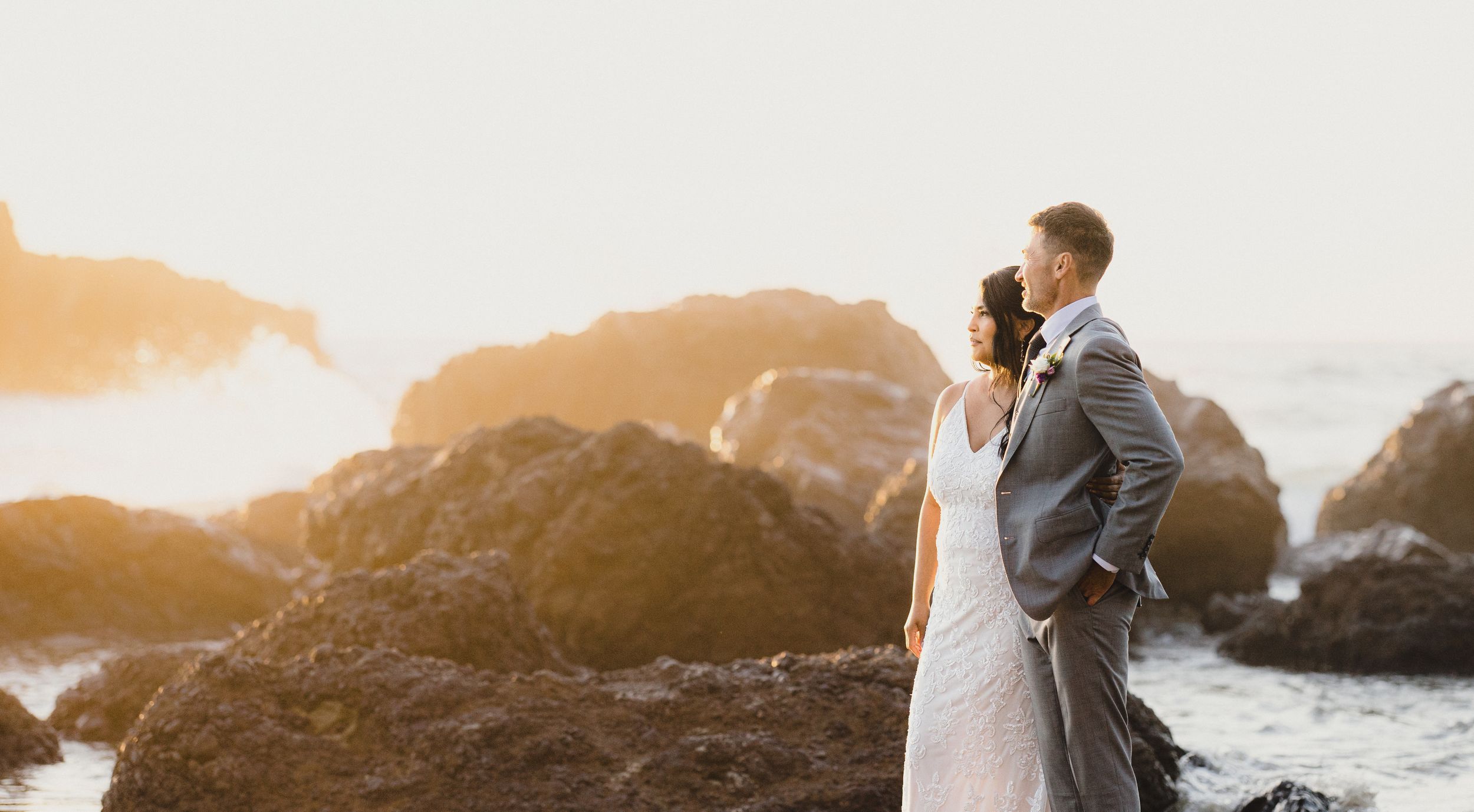 Bride and groom share a romantic moment on rocky coastal cliffs at golden sunset during their beach wedding.