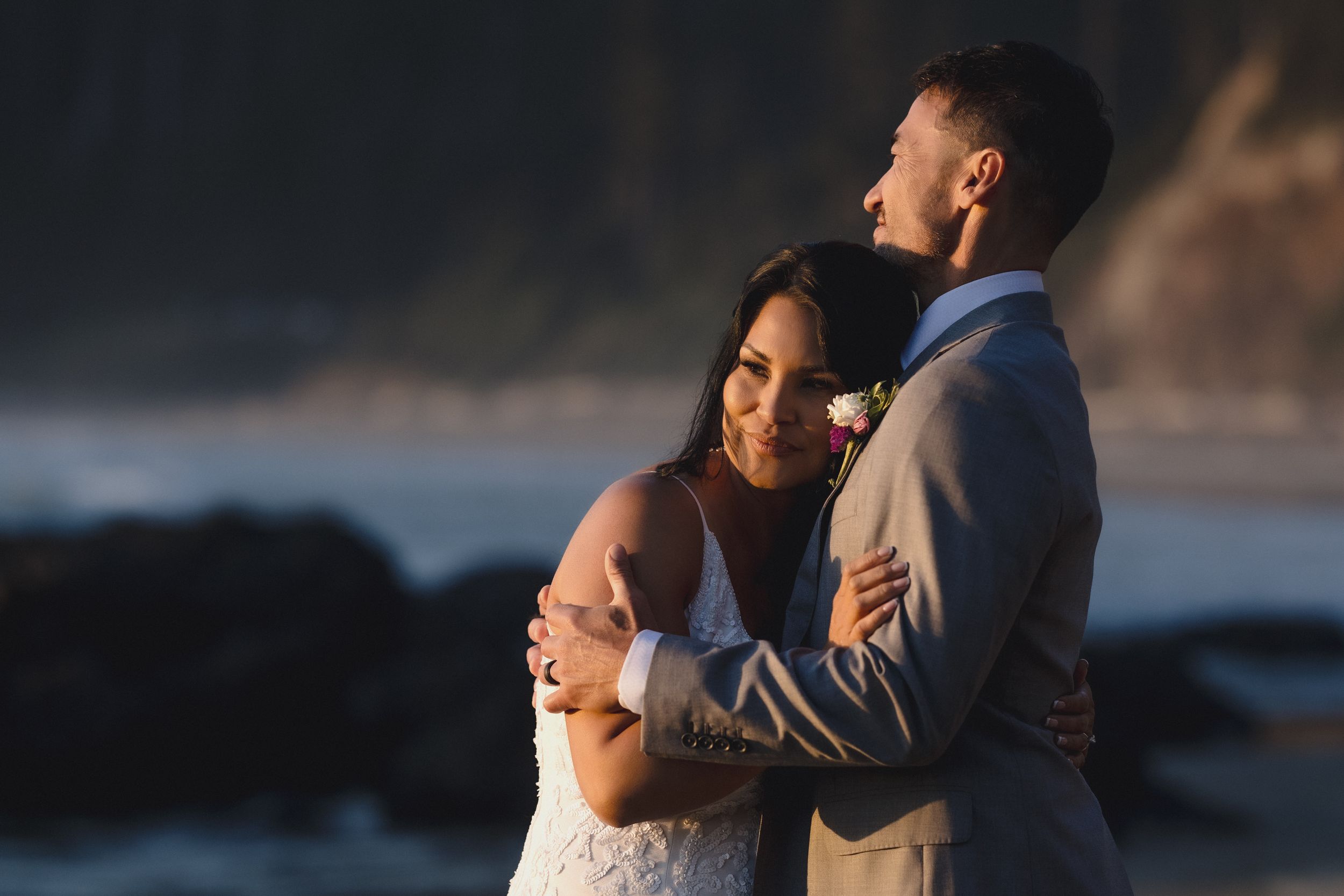 A couple embraces on a rocky beach at sunset, the bride in a lace gown and groom in a gray suit with boutonniere.