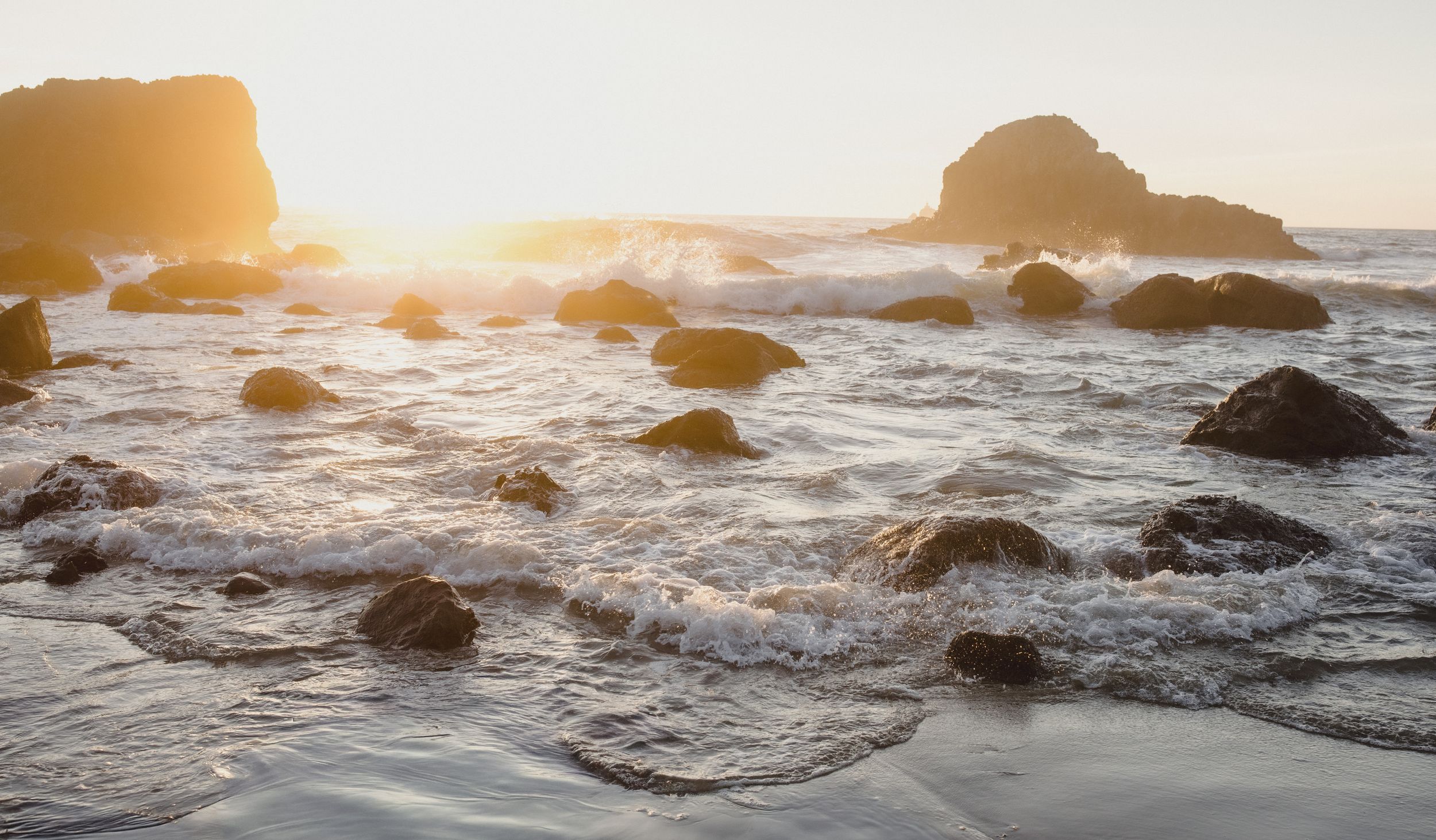 Golden sunset illuminates rocky shoreline with crashing ocean waves and sea stacks silhouetted against a warm glowing sky.