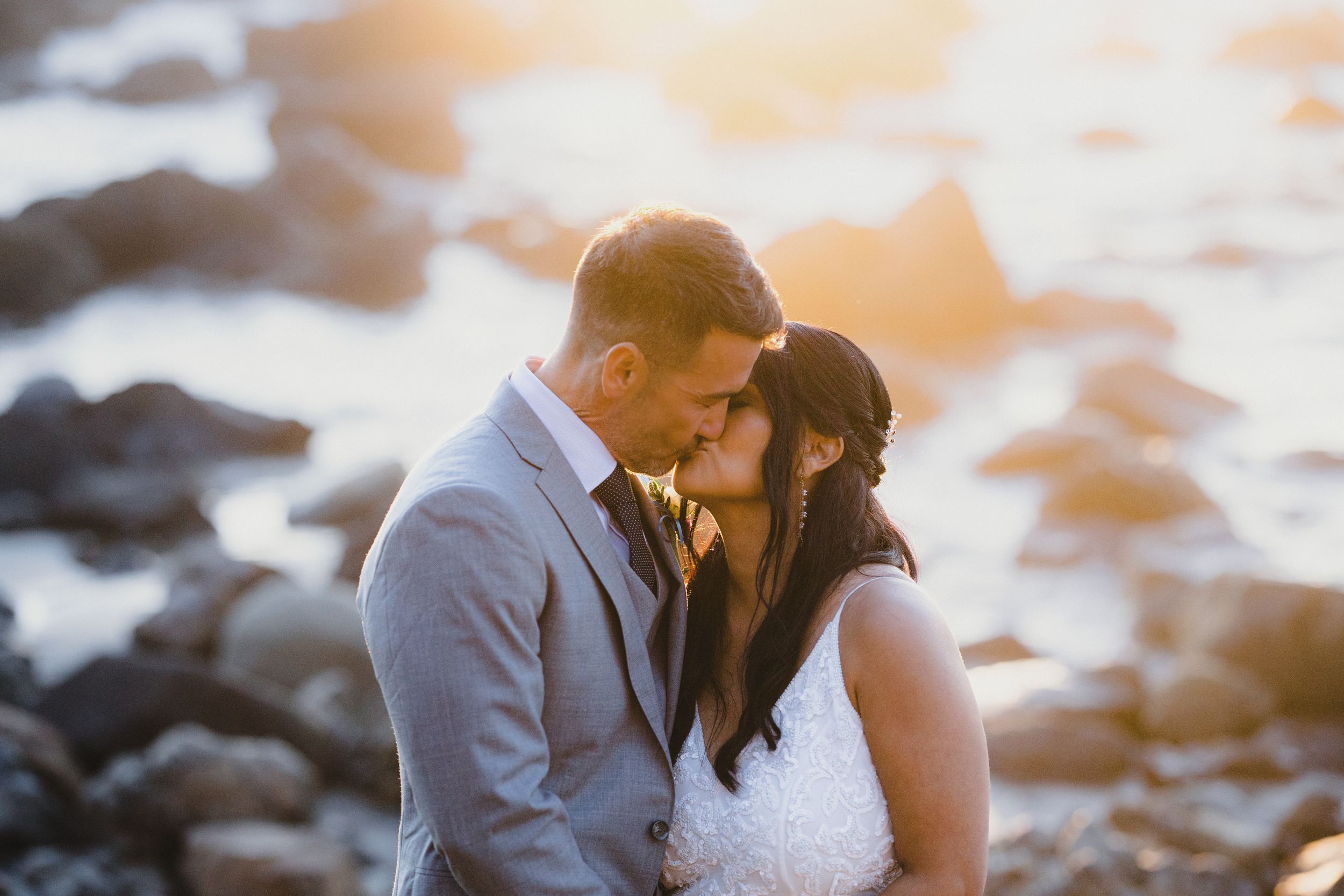 Bride and groom share a romantic moment near rocky shoreline at golden hour sunset.