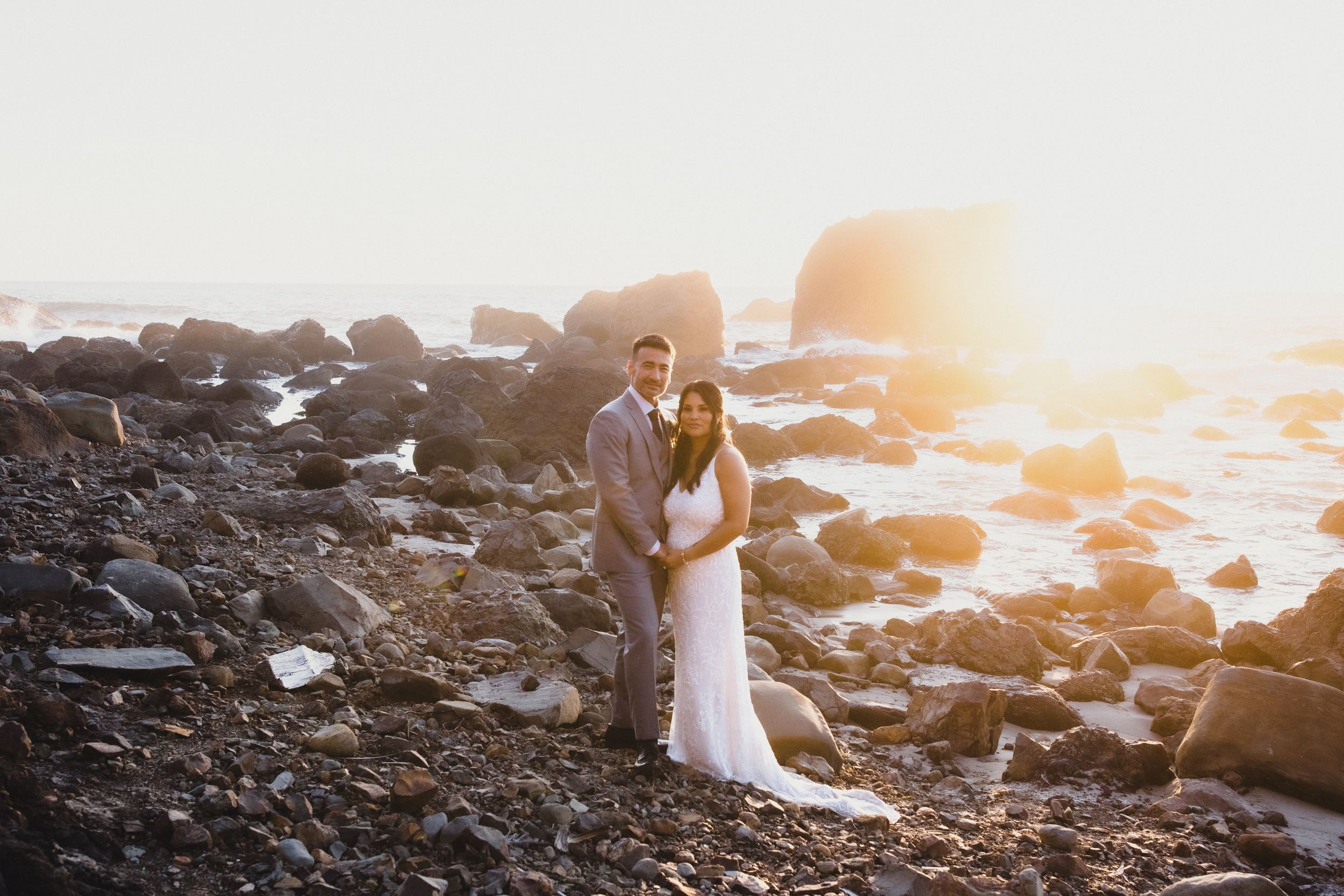 Bride and groom embrace on rocky beach at golden sunset, waves and boulders in background.