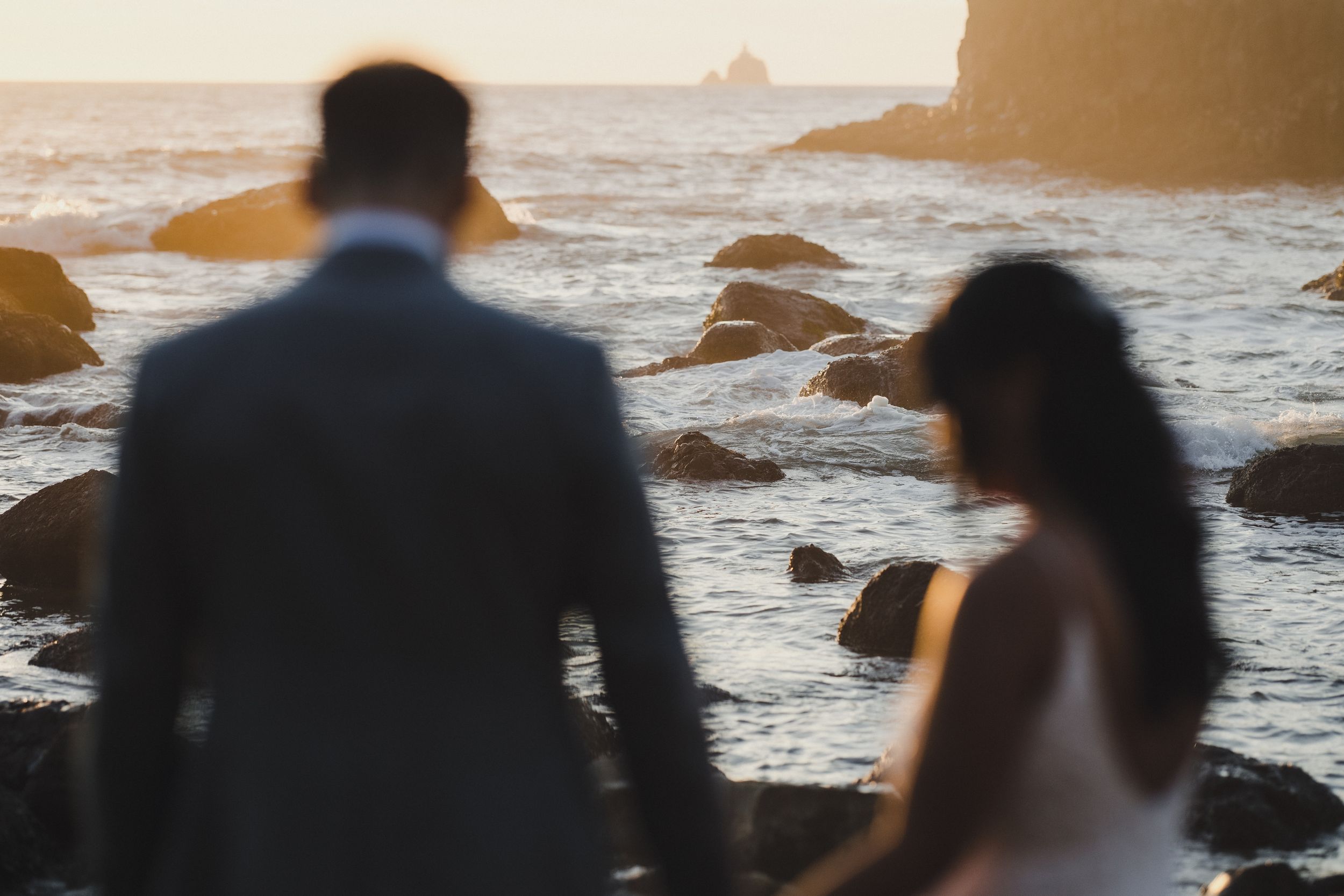 Couple in wedding attire standing on rocky coastal shore at golden sunset with ocean waves in background.