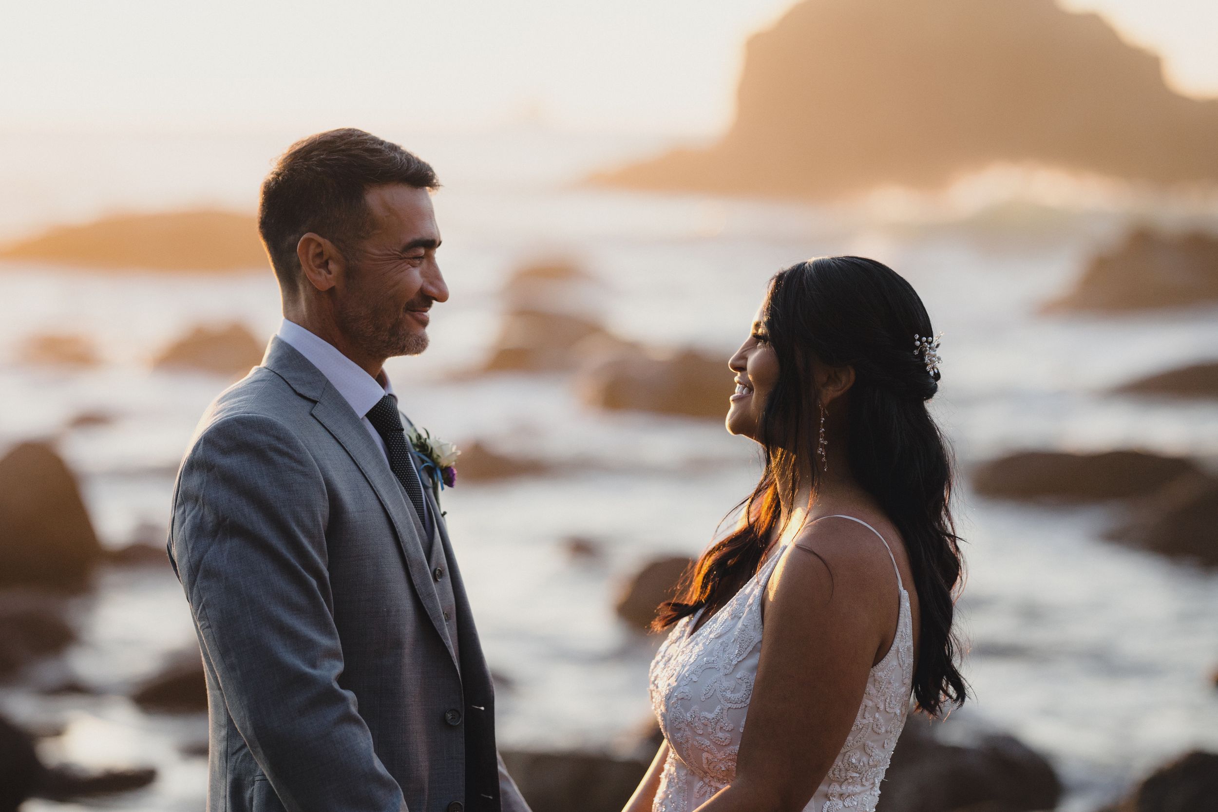 Couple in wedding attire sharing a romantic moment on rocky beach at golden sunset.
