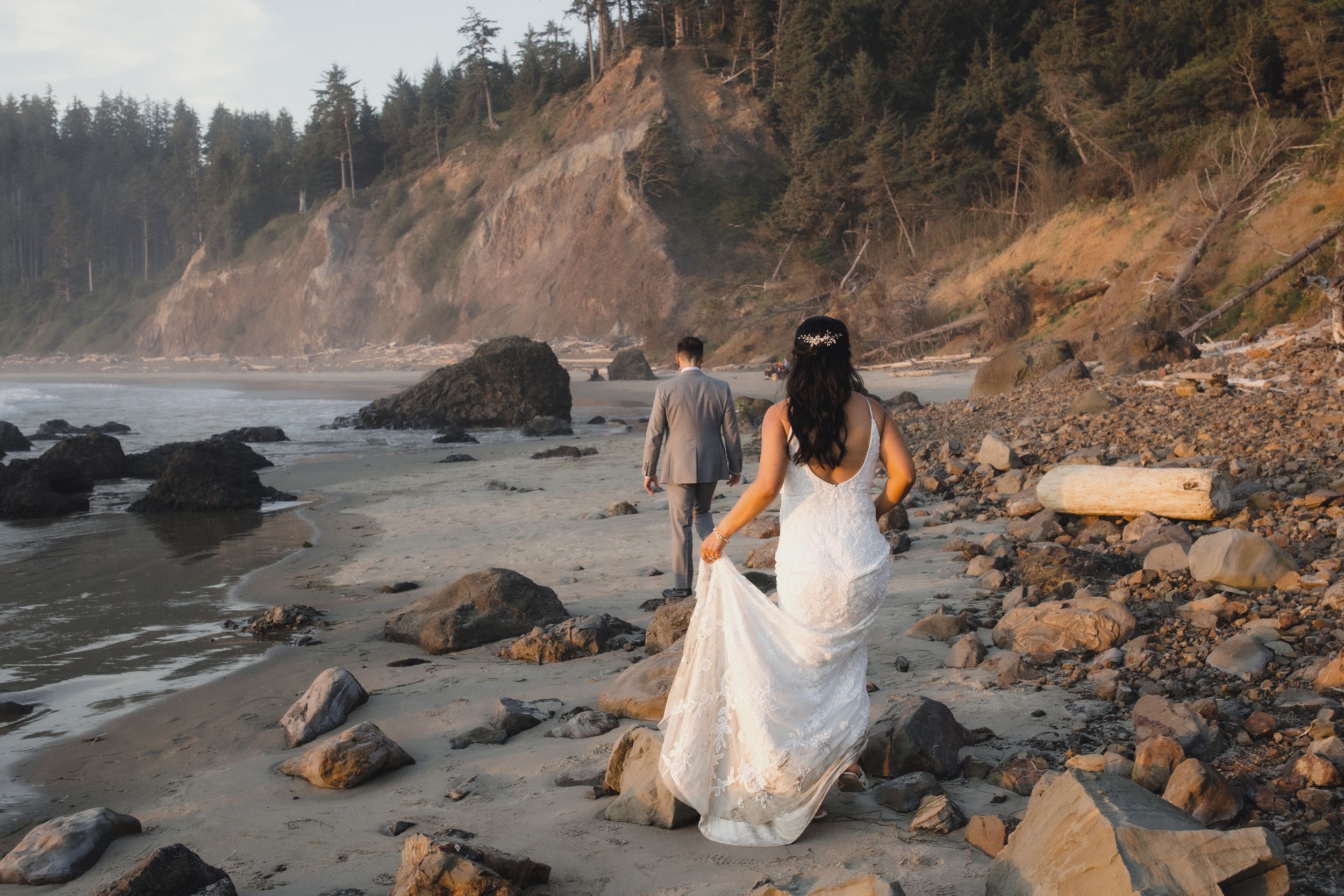 Bride and groom walking hand in hand on a rocky Pacific Northwest beach with dramatic cliffs and evergreen trees.