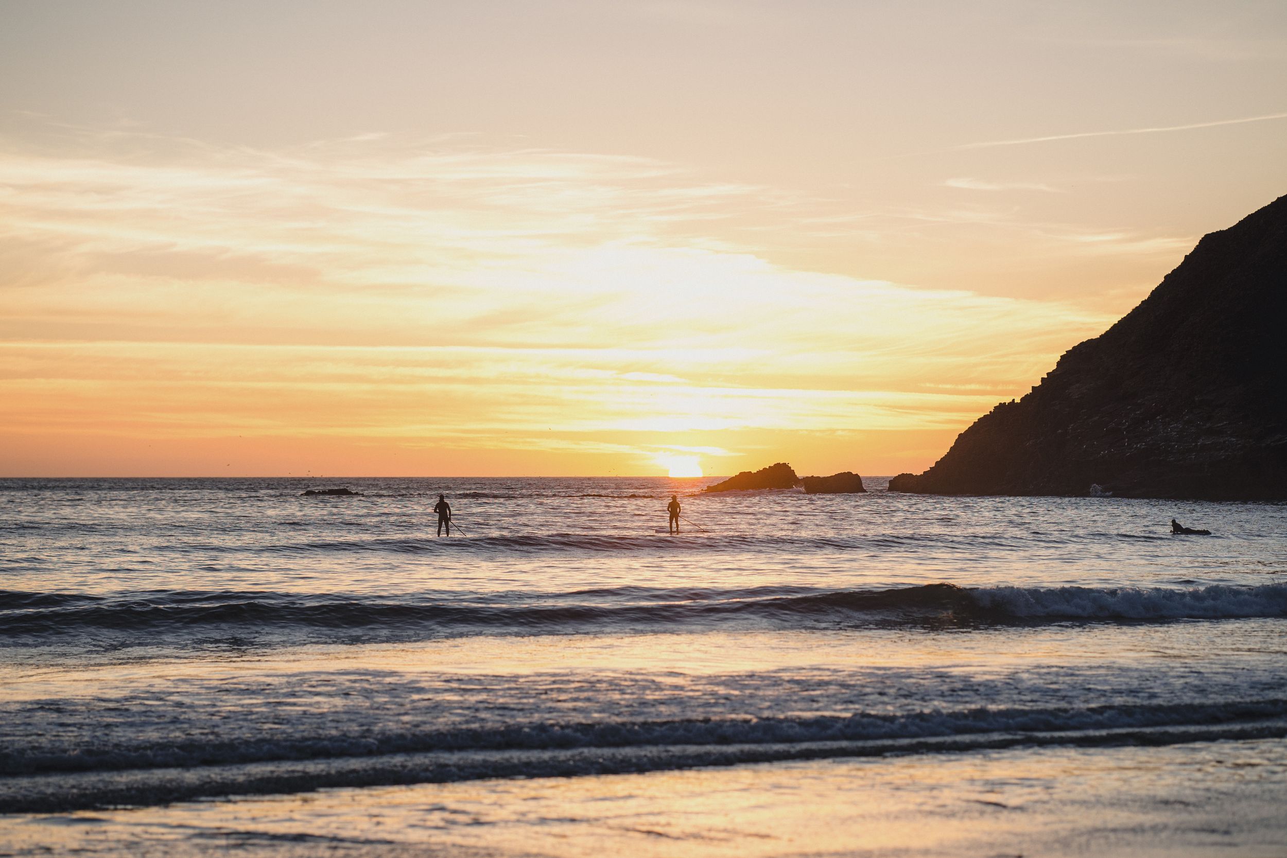 Silhouettes of surfers in ocean waves at stunning golden sunset with coastal hill backdrop.