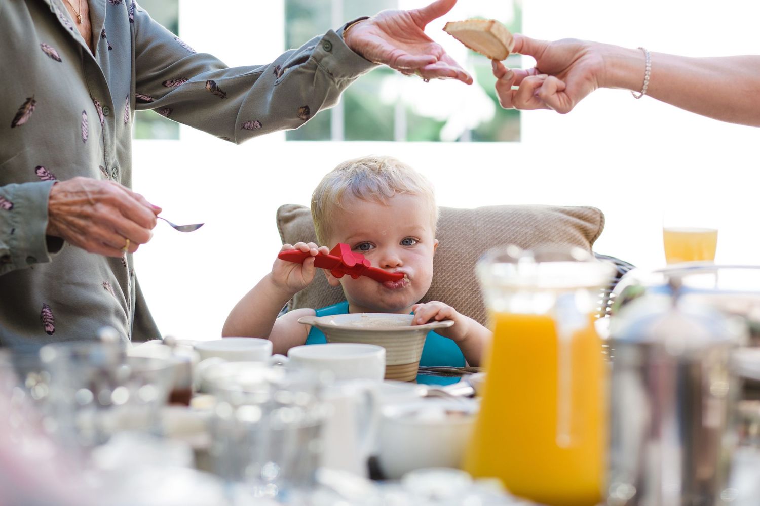 Toddler in high chair being fed by adults at a family breakfast table with juice and dishes.