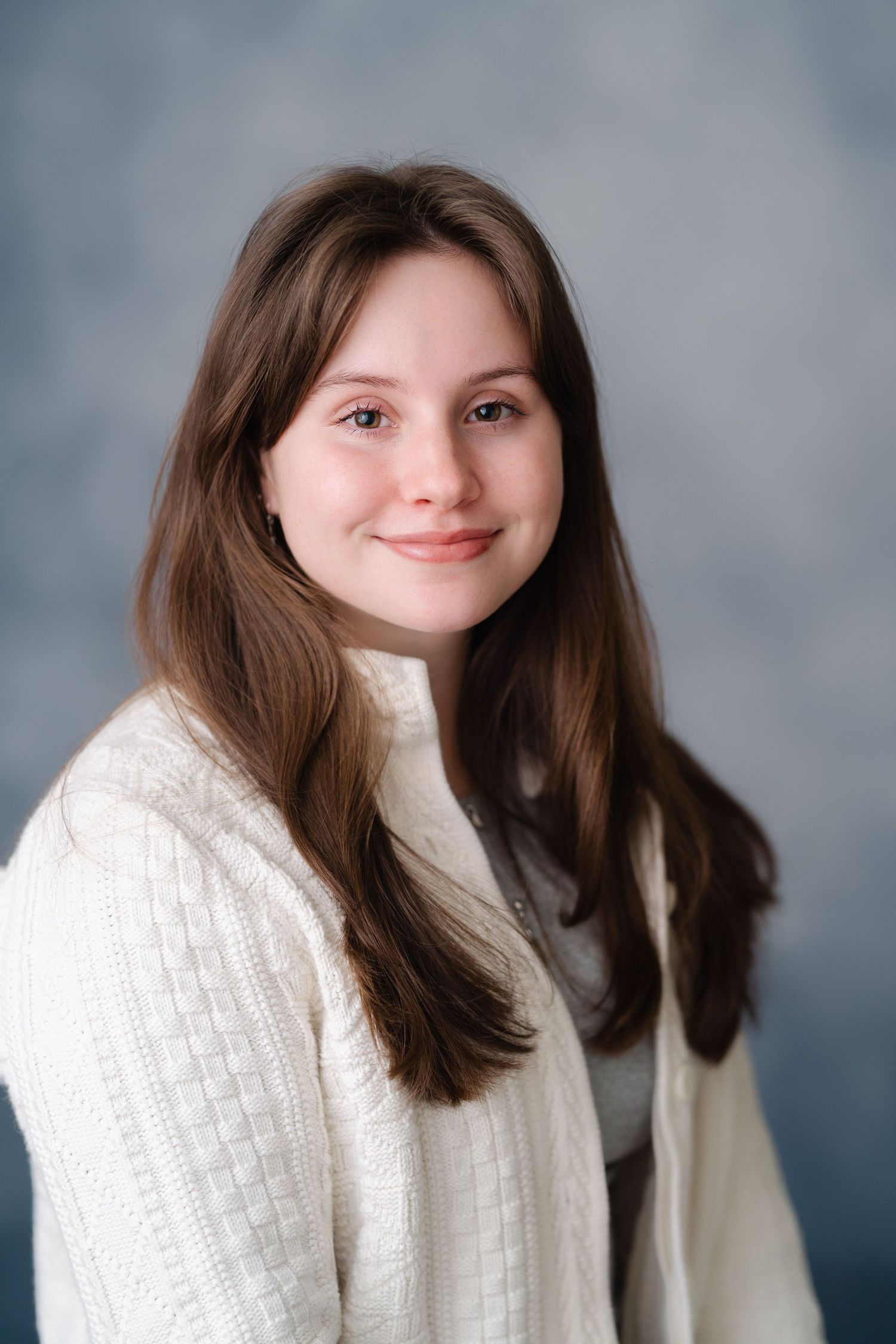 Young woman with long brown hair smiling, wearing a white knit sweater, posed against a soft blue studio background.