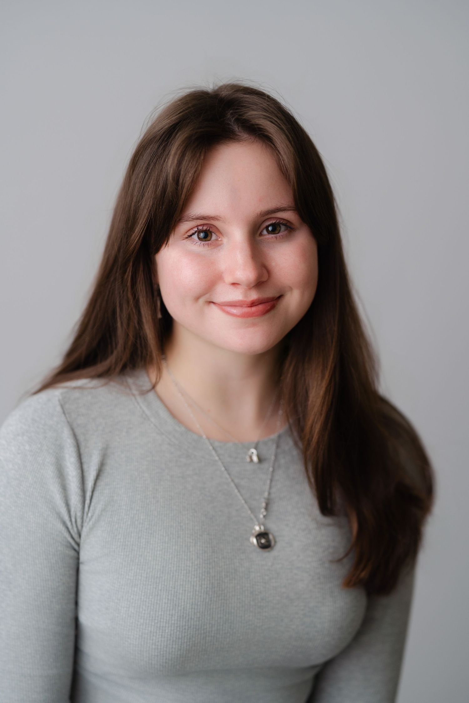 Young woman with long brown hair smiling, wearing a gray top and silver necklace, against a light gray background.