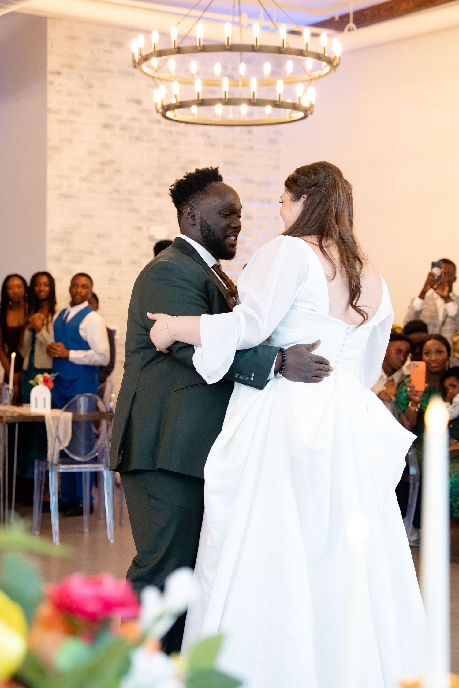 Bride and groom share their first dance under a chandelier at a wedding reception with guests watching.