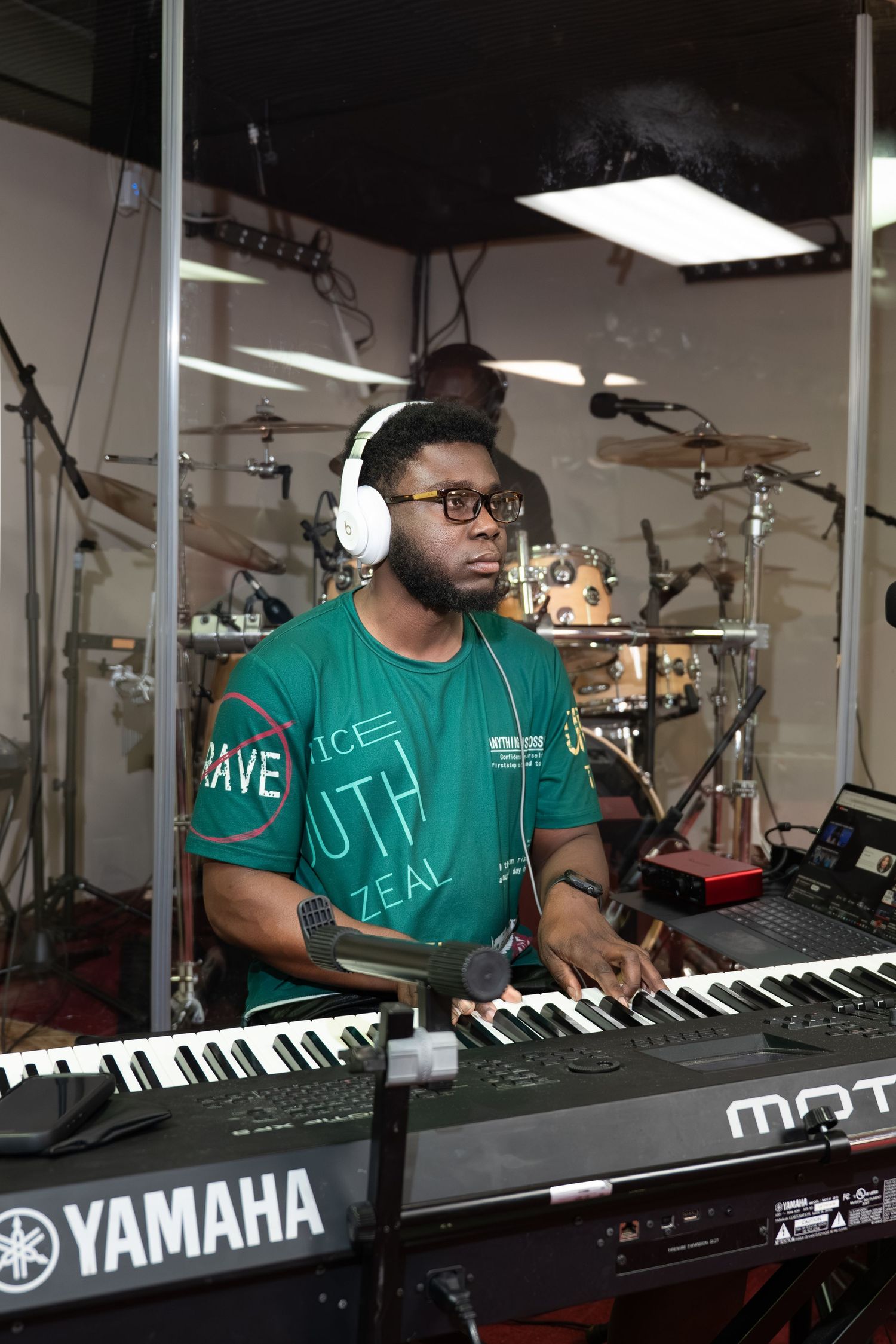 Man with headphones playing a Yamaha keyboard in a music studio with drums in the background.