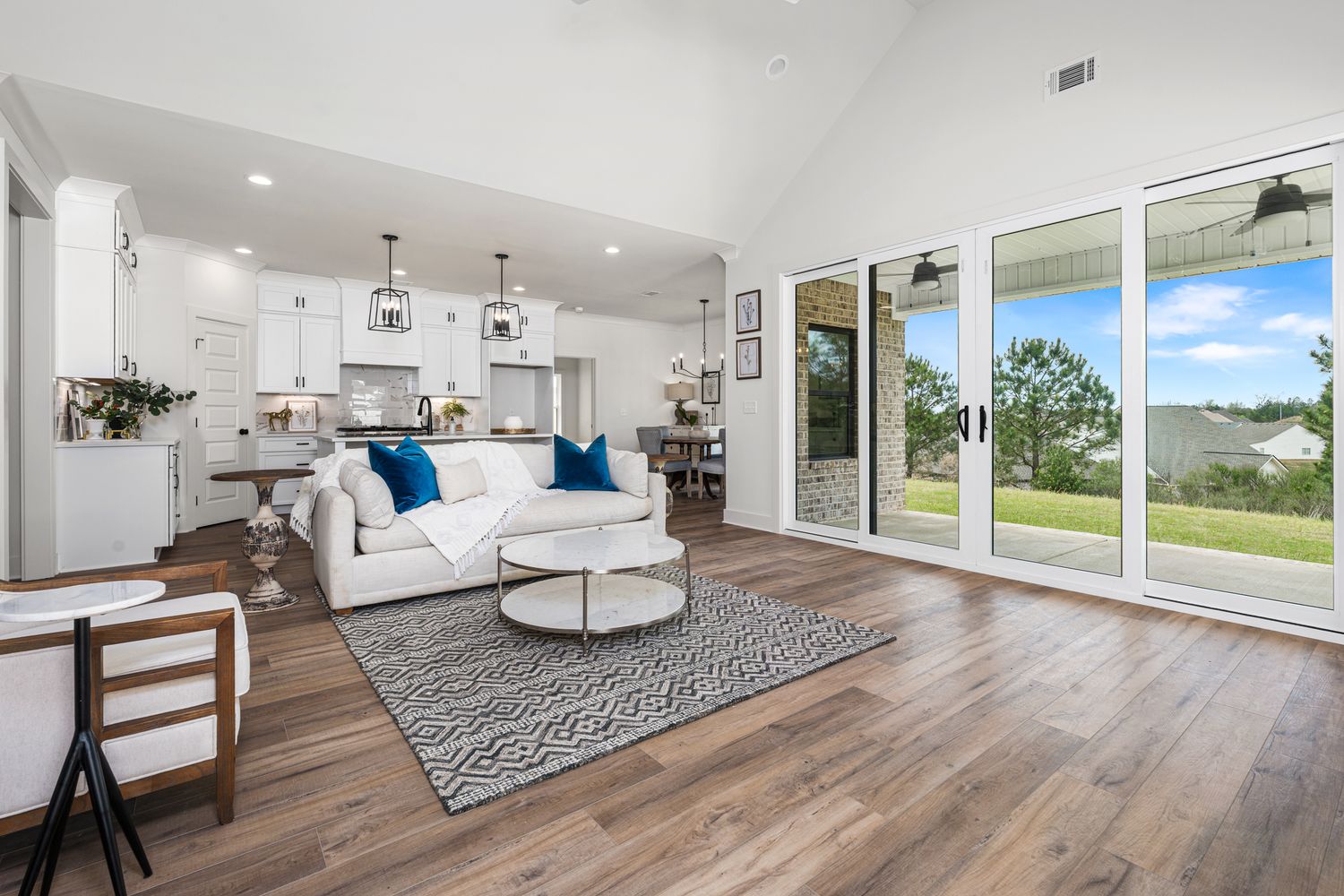 Bright open-concept living room with hardwood floors, white sofa, blue accents, high ceilings, and sliding glass doors to a scenic backyard.