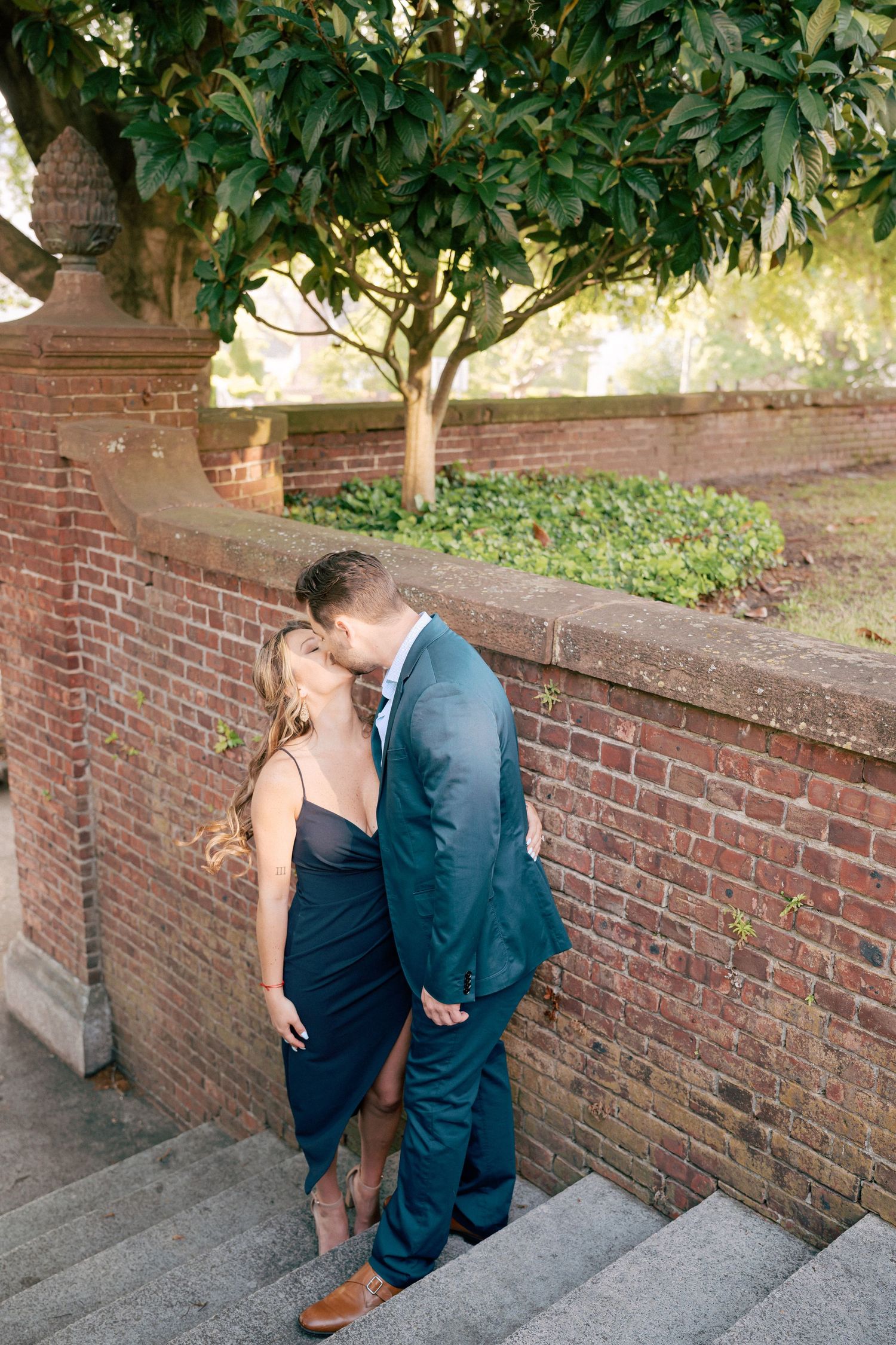 Couple sharing a romantic kiss on stone steps beside a brick wall with lush greenery, man in teal suit, woman in navy dress.