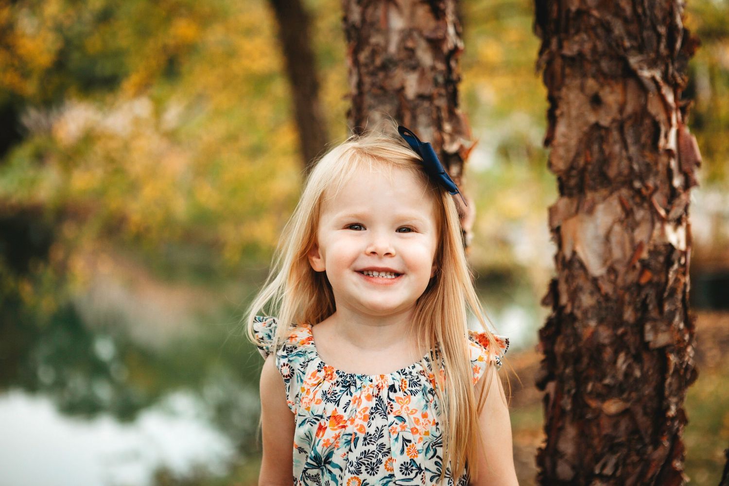 Smiling blonde girl with bow in hair wearing floral dress standing near trees in autumn park.