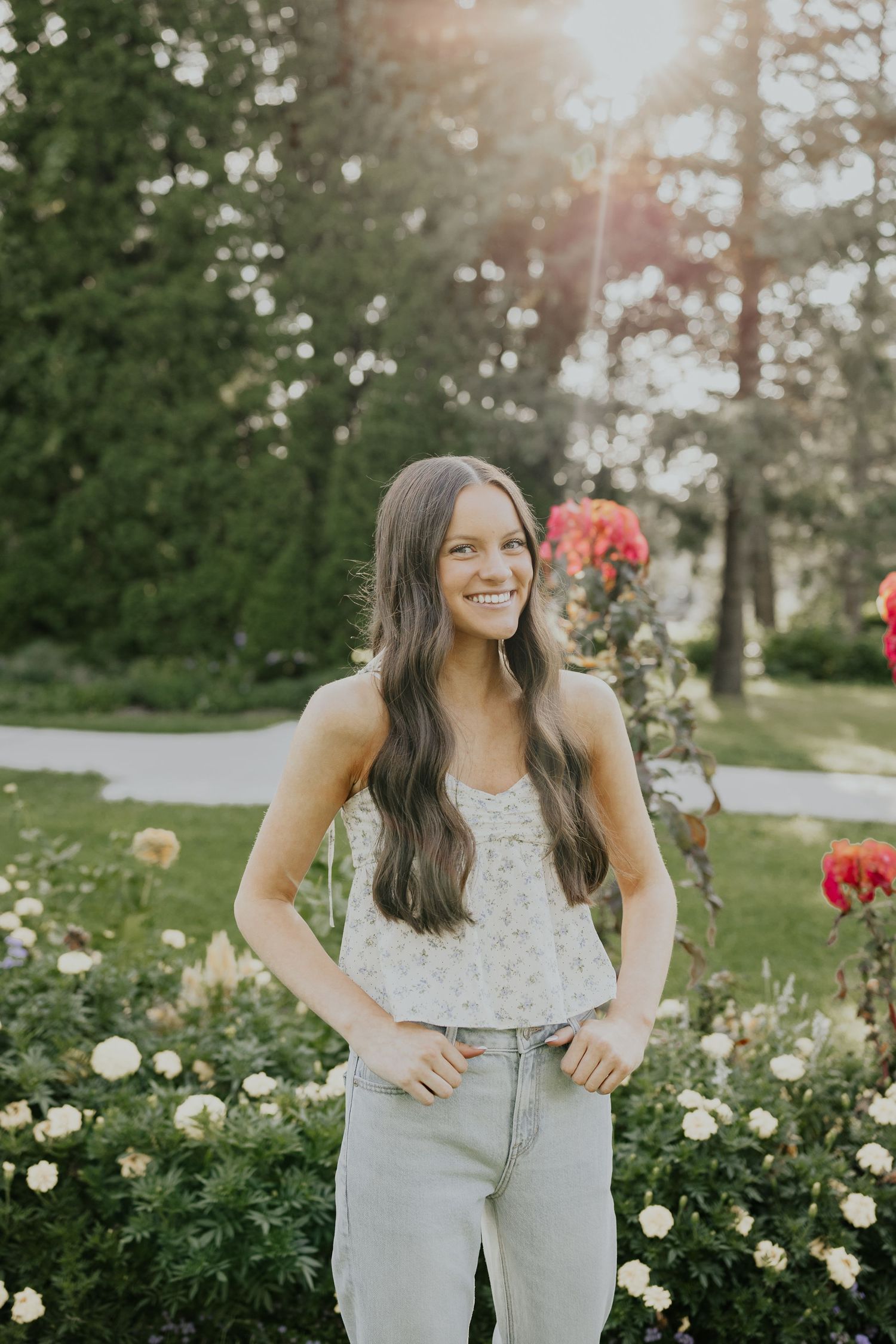 Smiling young woman in floral top and jeans standing in a sunny garden with colorful roses.