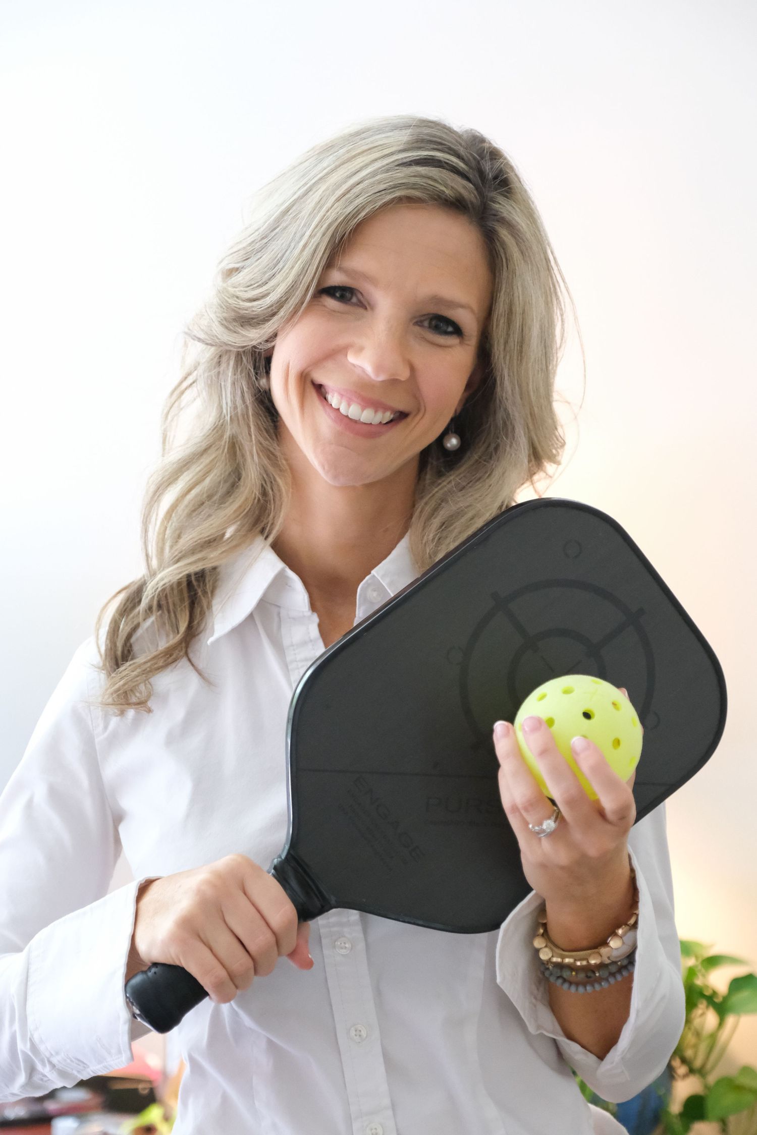 An environmental portrait of a woman with blonde hair wearing a white shirt holding a pickle ball paddle with a yellow ball.