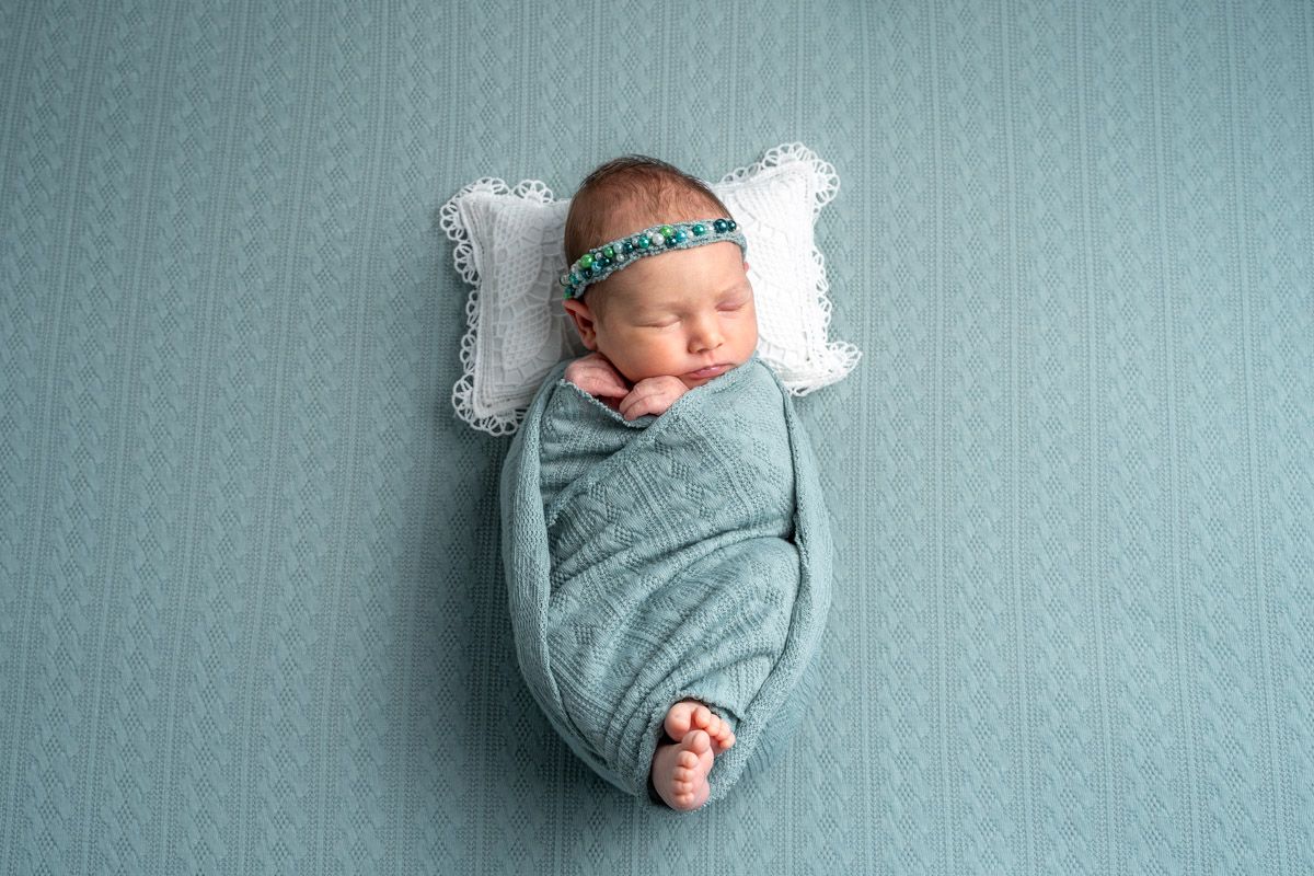 Newborn baby swaddled in teal knit wrap with floral headband, sleeping peacefully on white pillow against teal background.