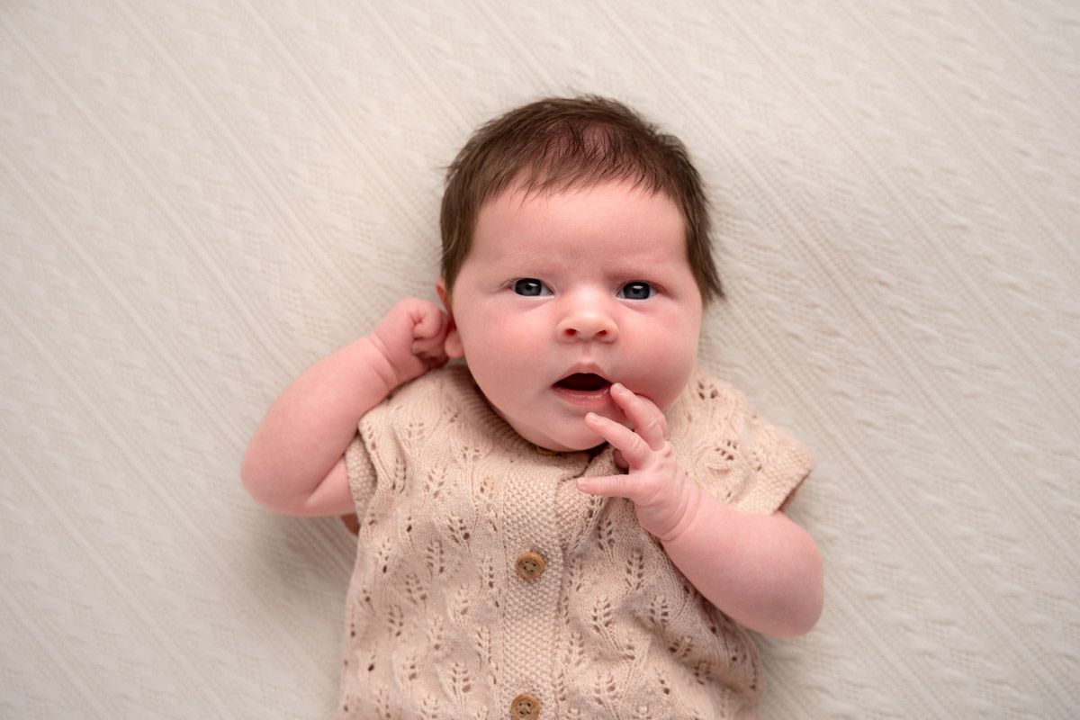 Newborn baby in a beige knit outfit lying on a white blanket, looking up with wide eyes and tiny hands raised.