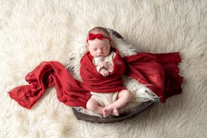 Newborn baby girl wrapped in red knit swaddle with bow headband, sleeping peacefully in wooden bowl on white fur.